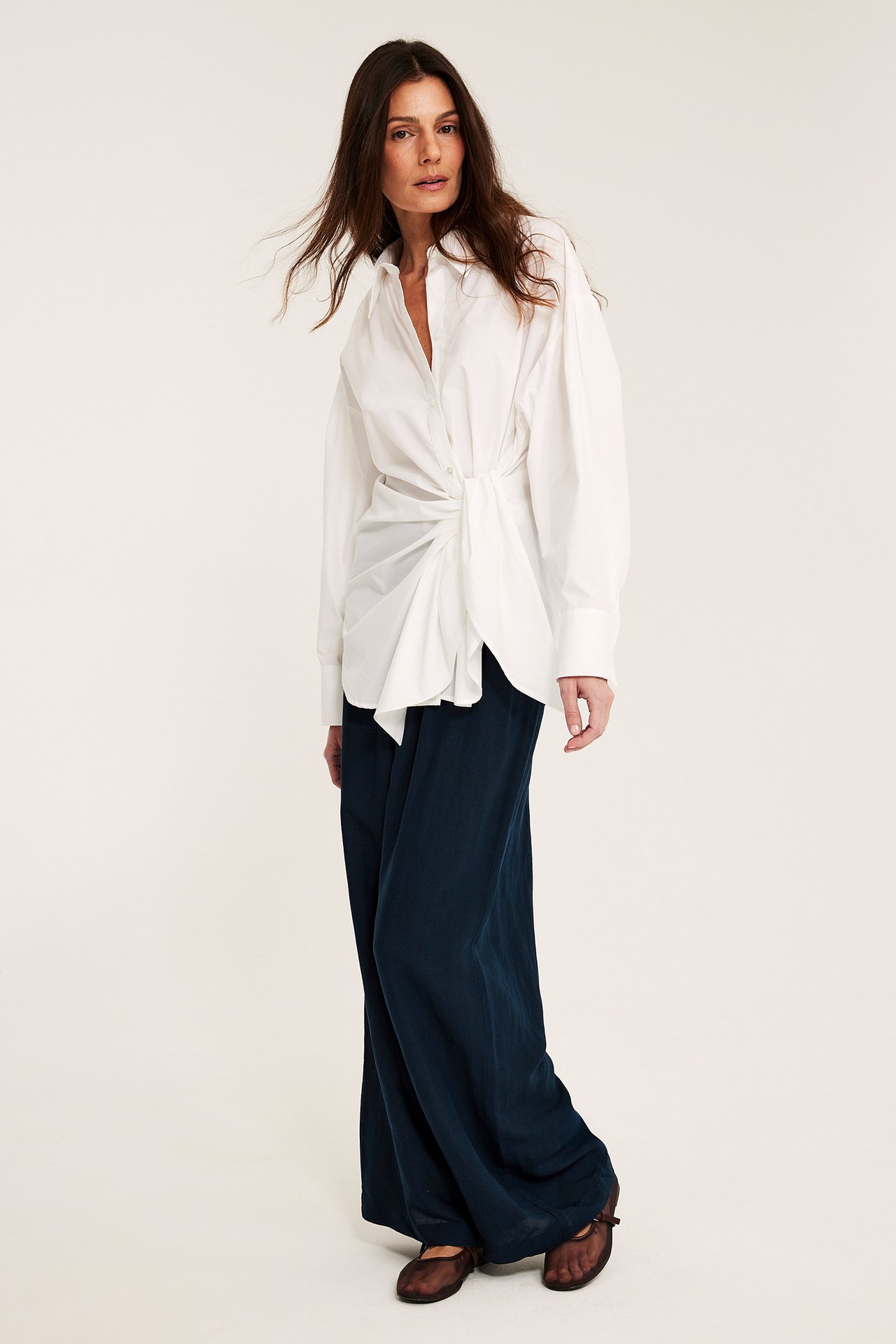 Widok z przodu: kobieta w ciemnoniebieskich szerokich spodniach lnianych, białej wiązanej koszuli i brązowych butach.