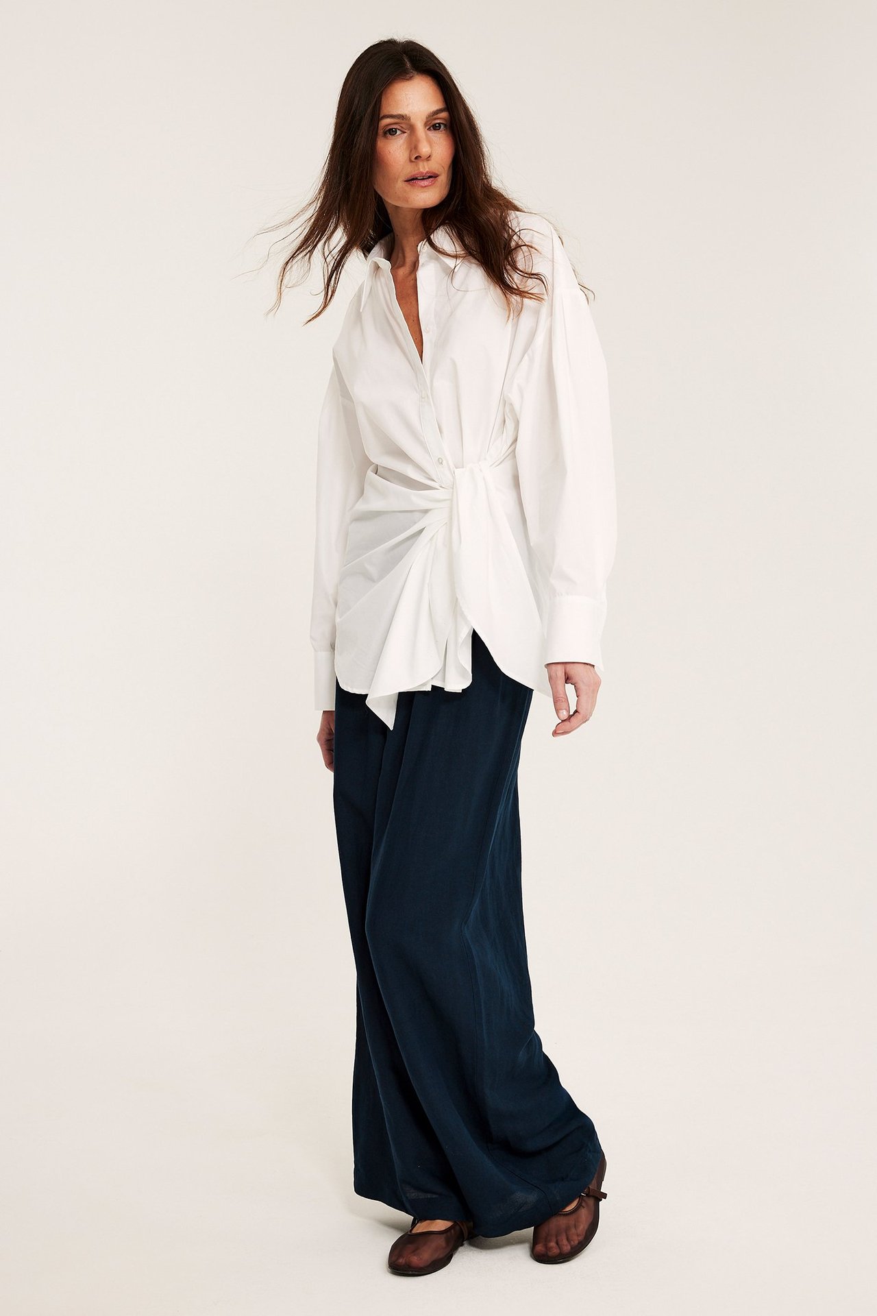 Frontbilde av kvinne i mørkeblå vide linblandingsbukser, hvit knyttet skjorte og brune sko.