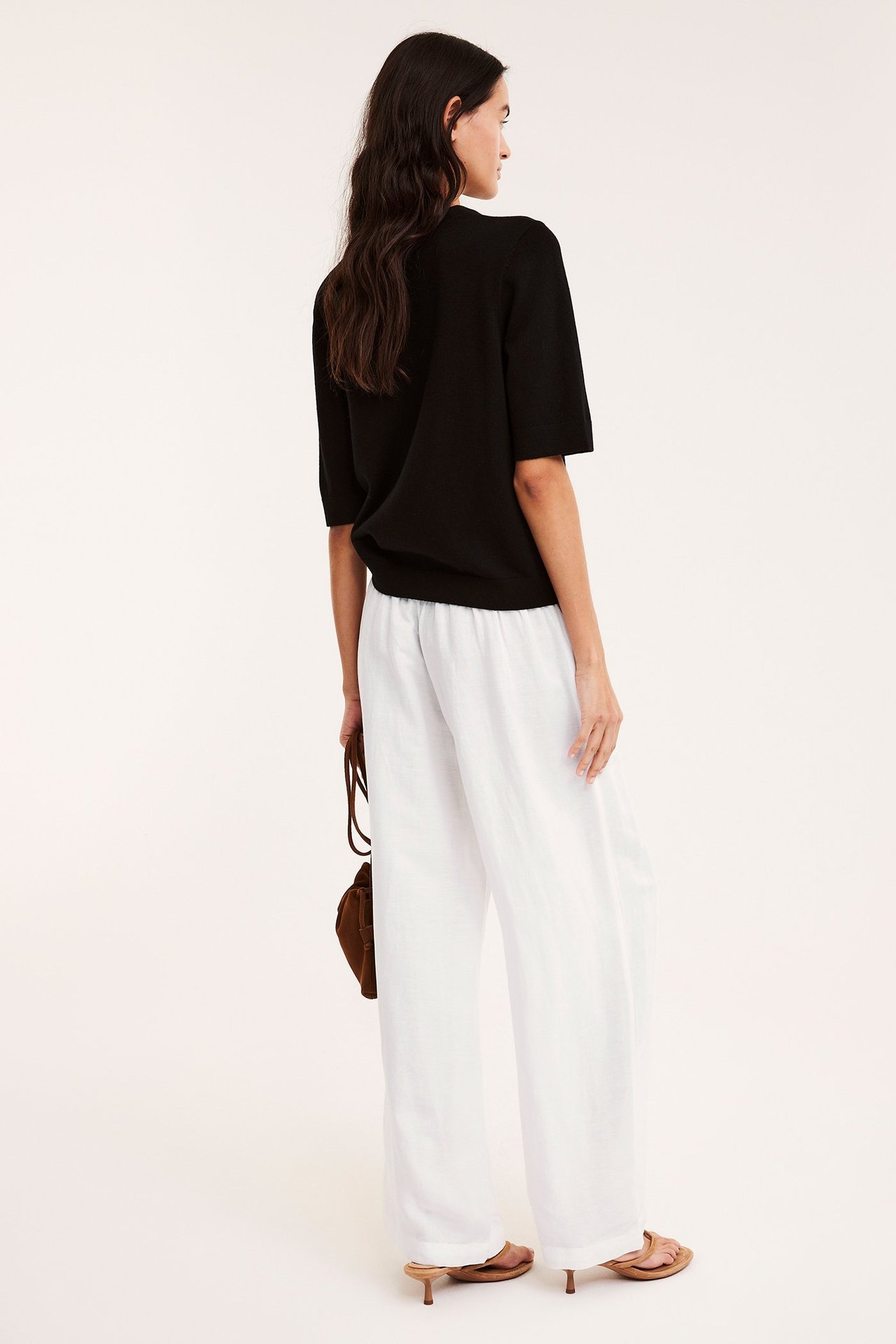 Nainen valkoisissa leveissä pellavahousuissa smock-vyötäröllä, musta toppi, ruskea laukku ja sandaalit.