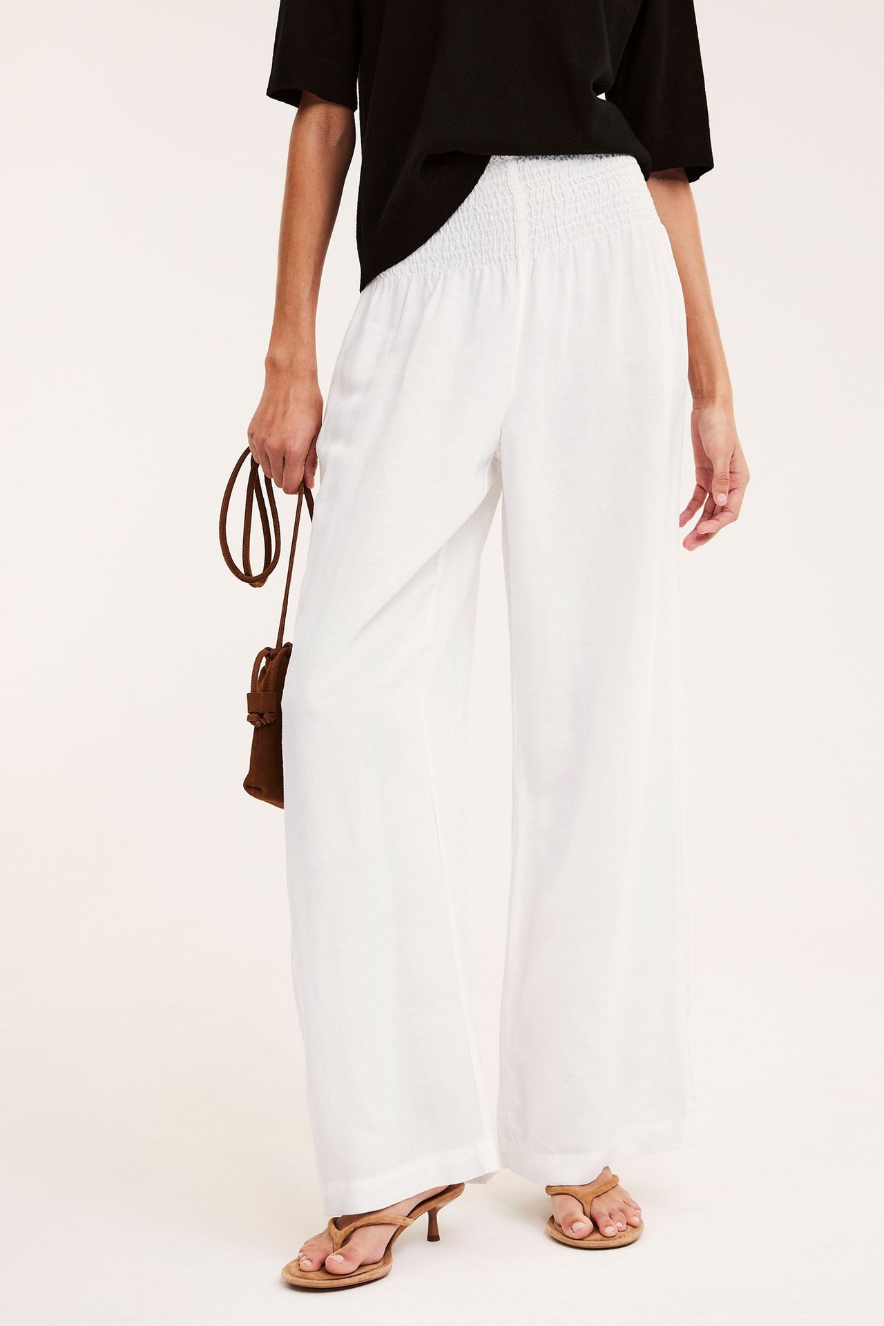 Kobieta w białych szerokich spodniach z lnu ze marszczoną talią, czarny top, brązowe sandały, trzyma brązową torebkę.