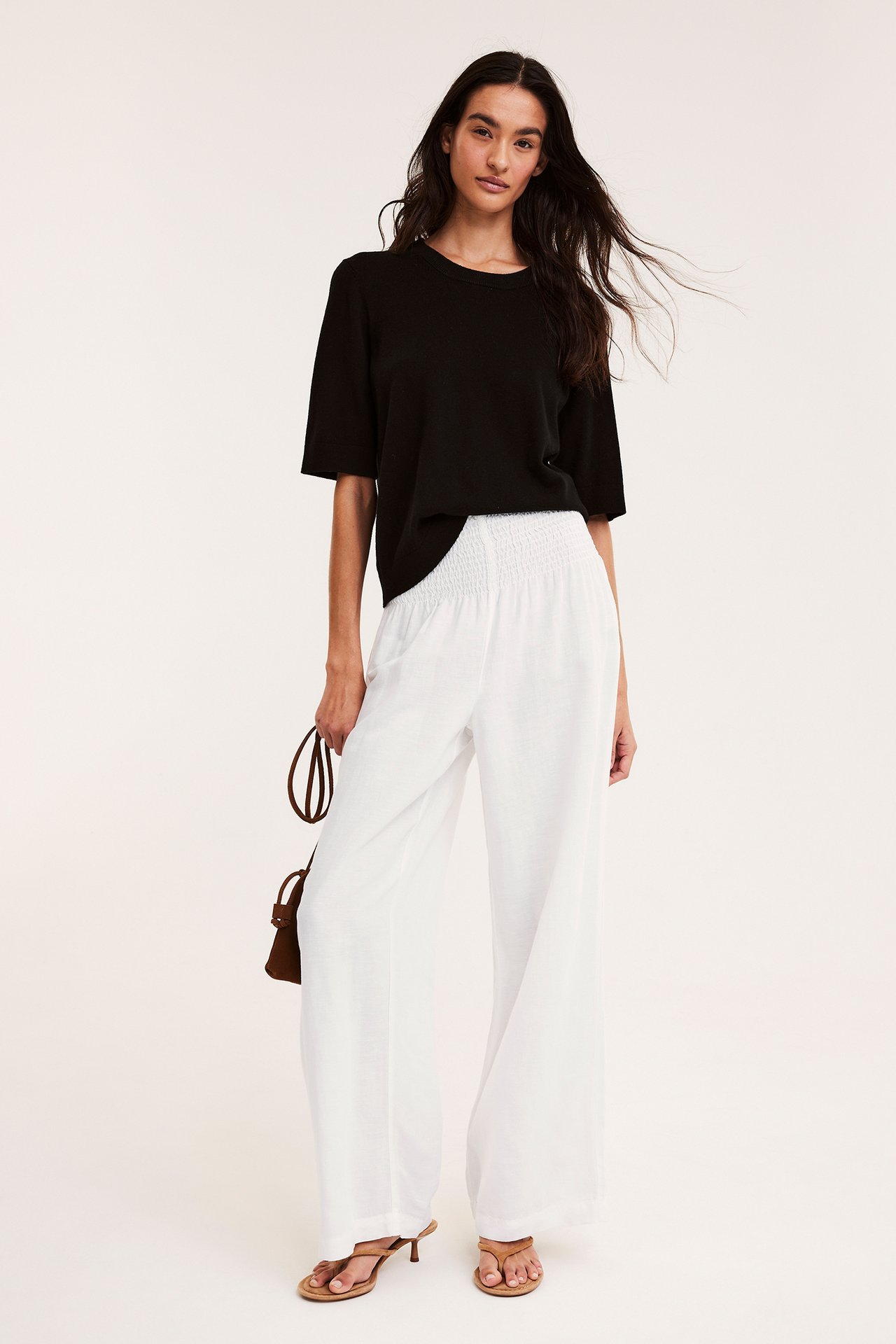 Kobieta w białych, szerokich spodniach z lnu ze marszczoną talią, czarny top, brązowe sandały, trzyma torebkę.