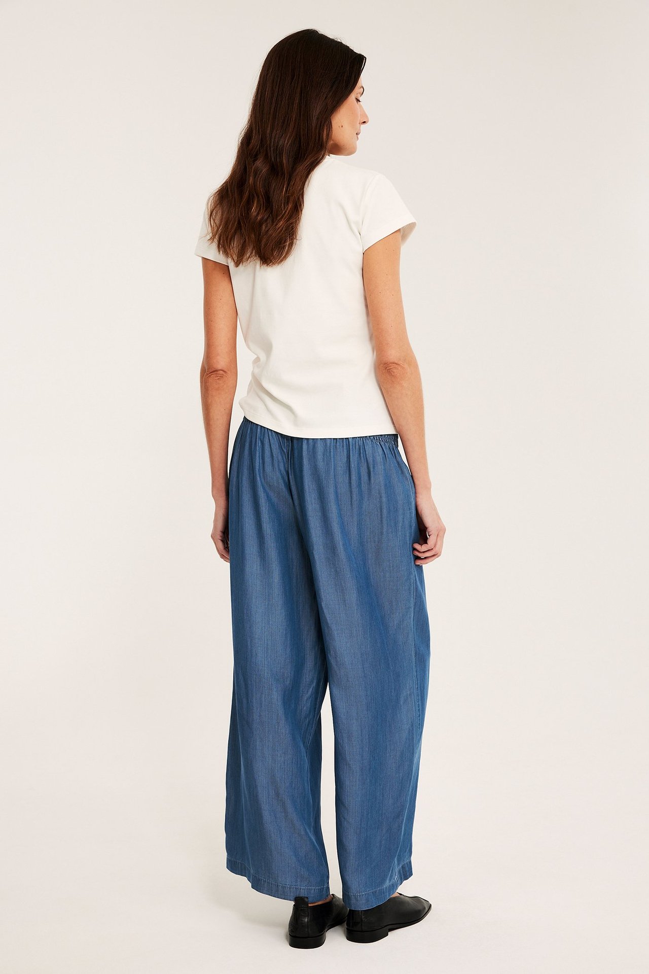 Takakuva: nainen yllään vaaleansiniset lyocell-housut, smokkiryppyvyötärö, valkoinen t-paita ja mustat kengät.