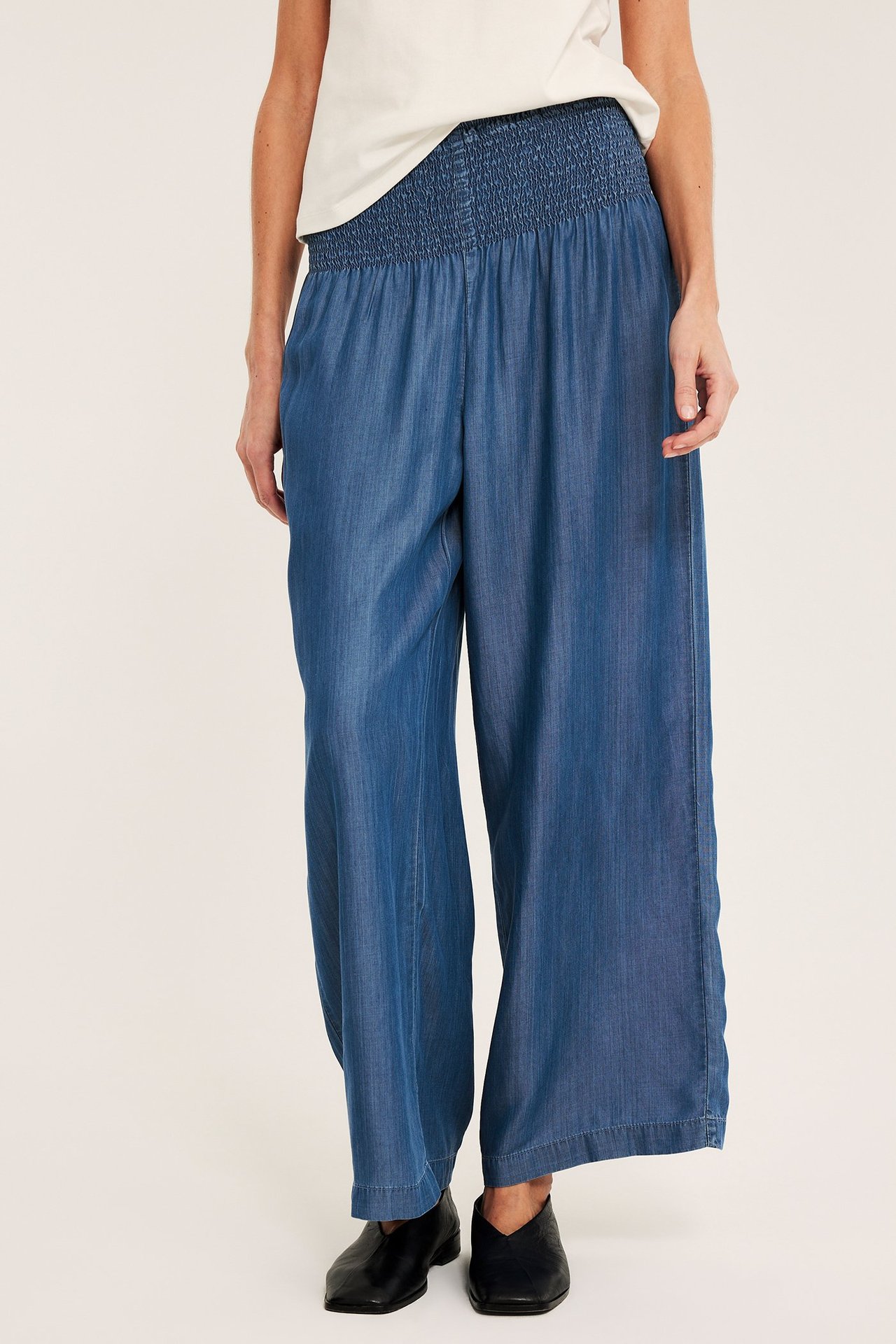 Edestä: nainen vaaleansinisissä, leveälahkeisissa lyocell-housuissa smokkivyötäröllä, valkoisessa topissa ja mustissa kengissä.