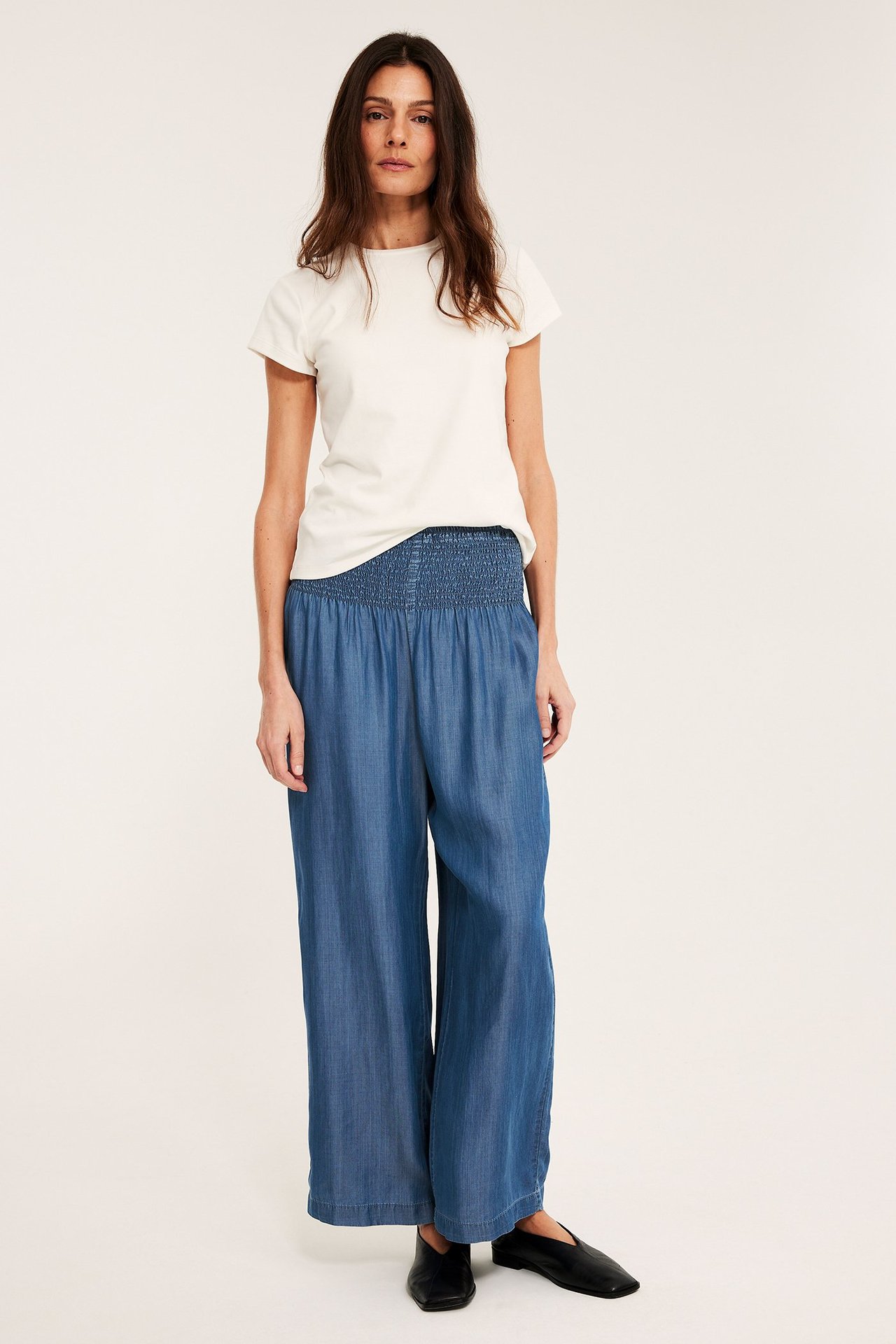 Framifrån: Kvinna i ljusblå lyocellbyxor med vida ben och smockmidja, krämvit t-shirt, svarta flats.