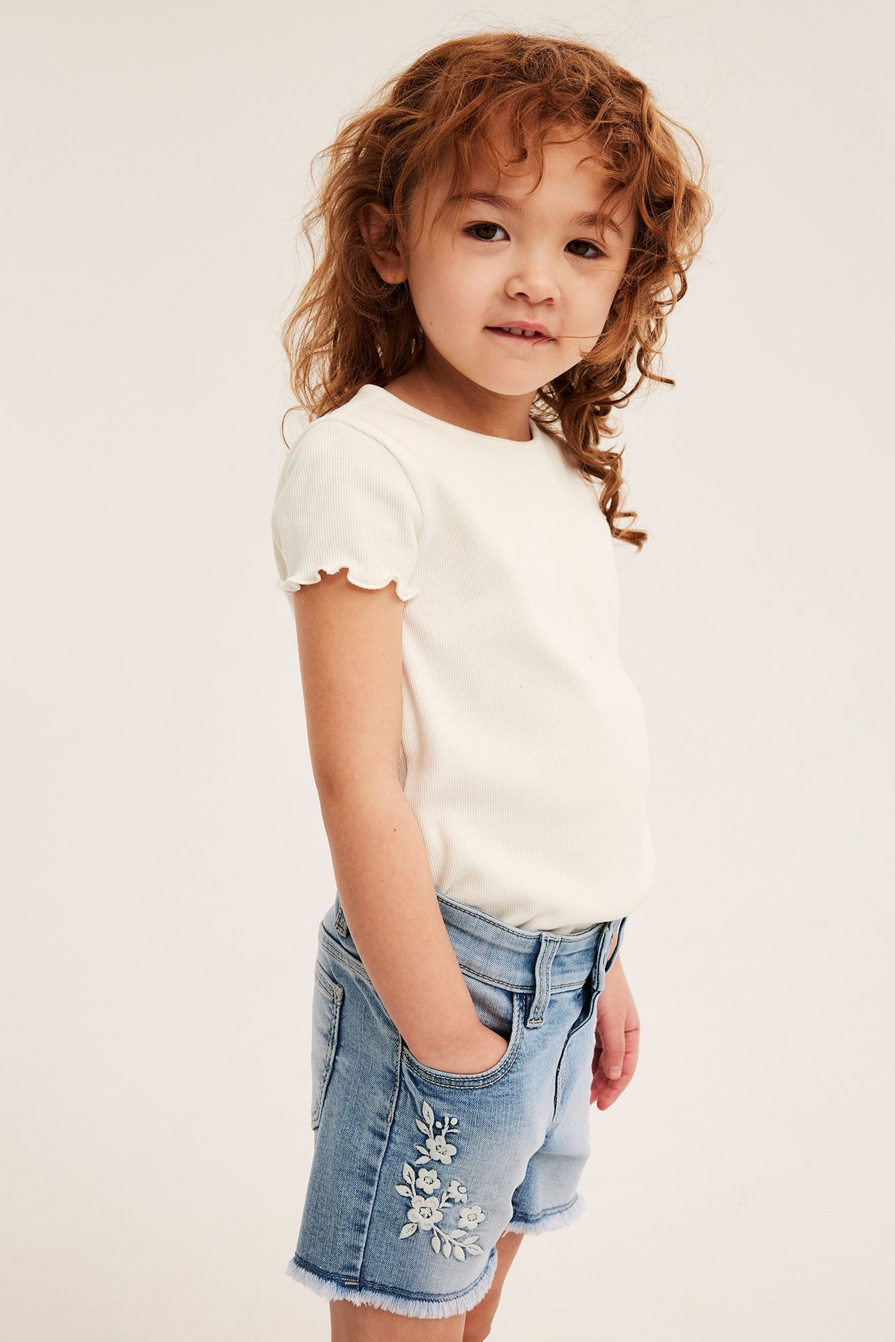 Etusivukuva: Lapsi yllään vaaleansiniset farkkushortsit valkoisella kukkakirjonnalla ja valkoinen ribbipaita.