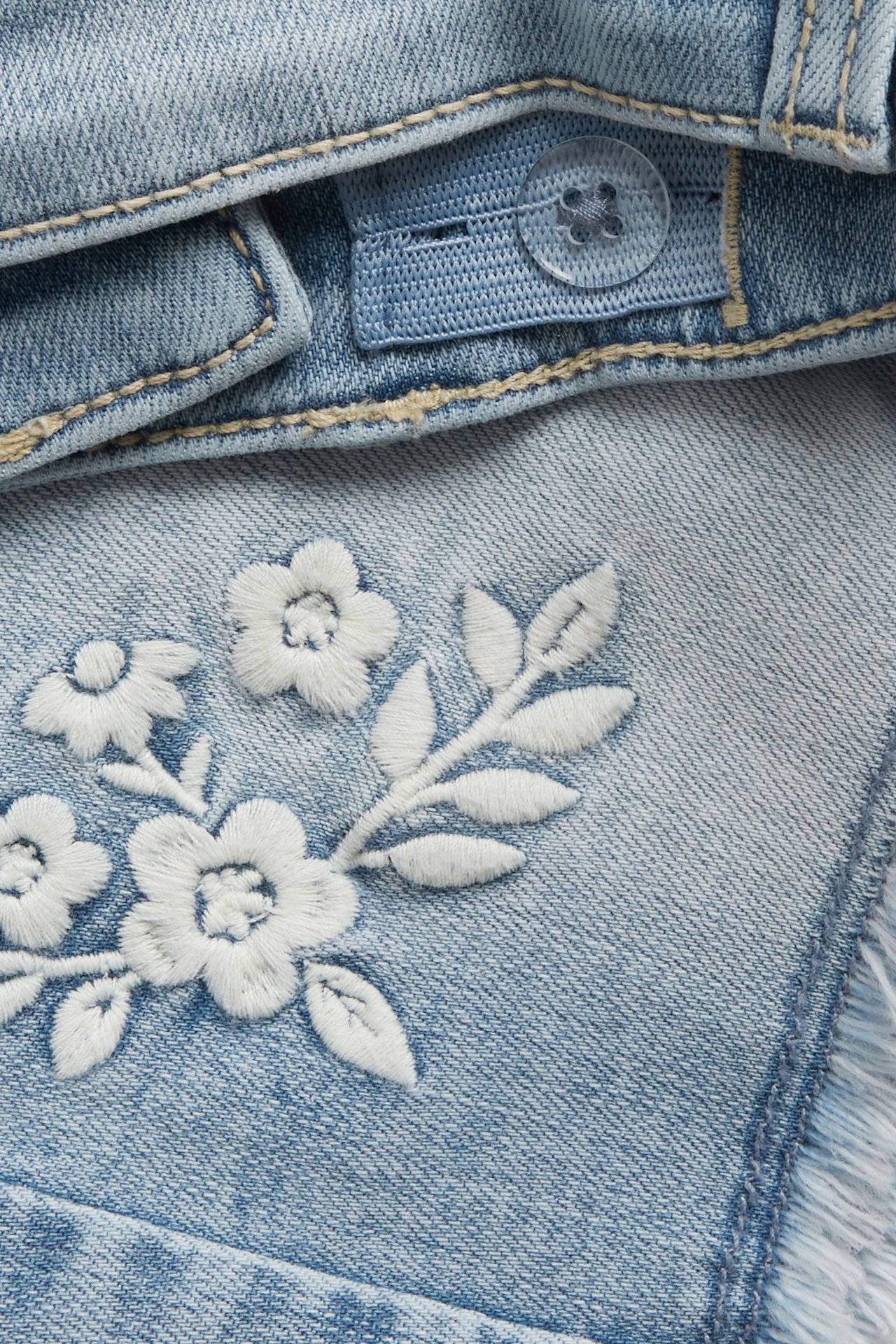 Zbliżenie jasnoniebieskich elastycznych szortów jeansowych z białym haftem kwiatowym i regulowanym guzikiem w pasie.