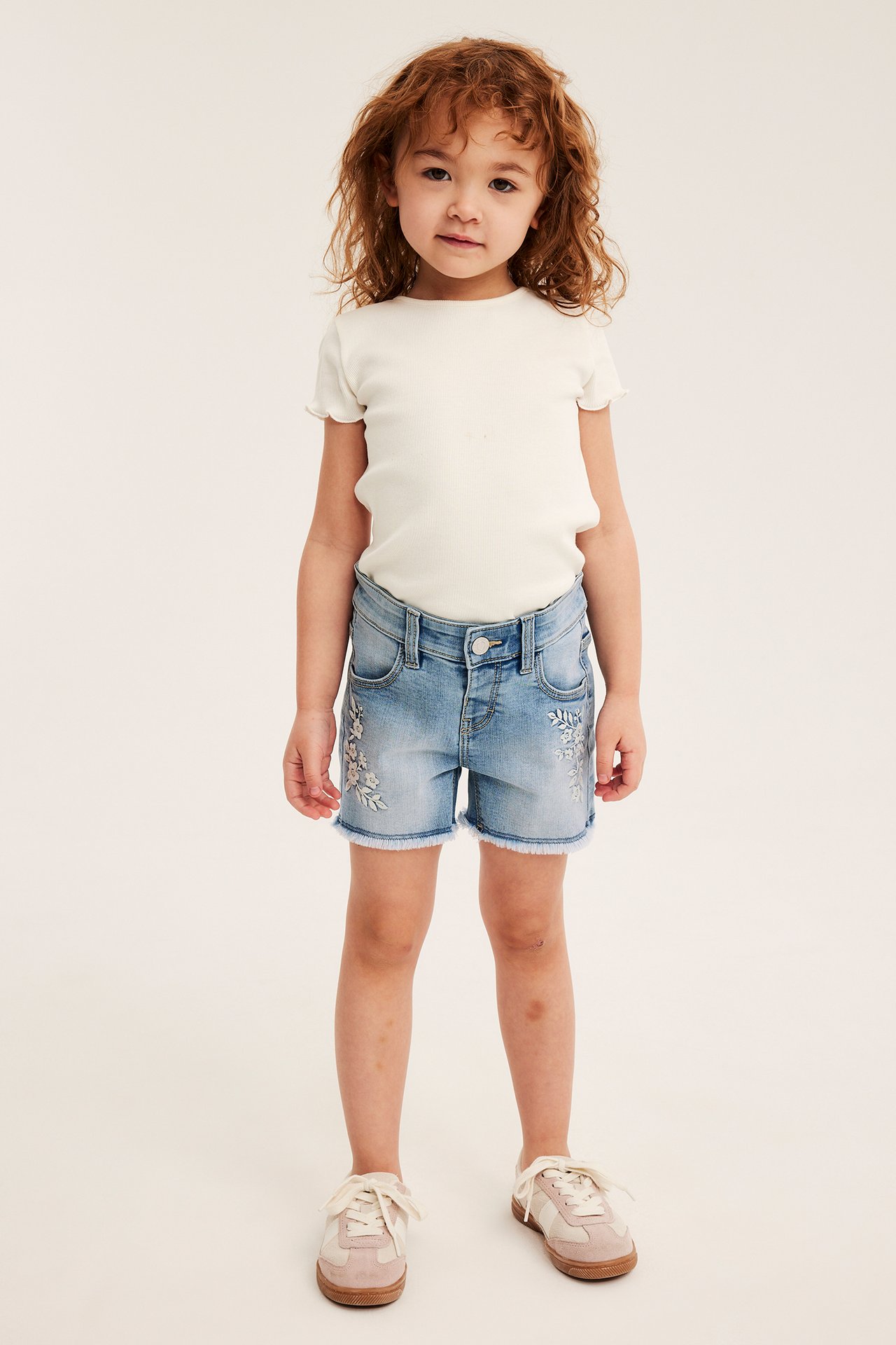 Framifrån: Kid i ljusblå broderade jeansshorts, en ribbad benvit t-shirt och beiga sneakers.