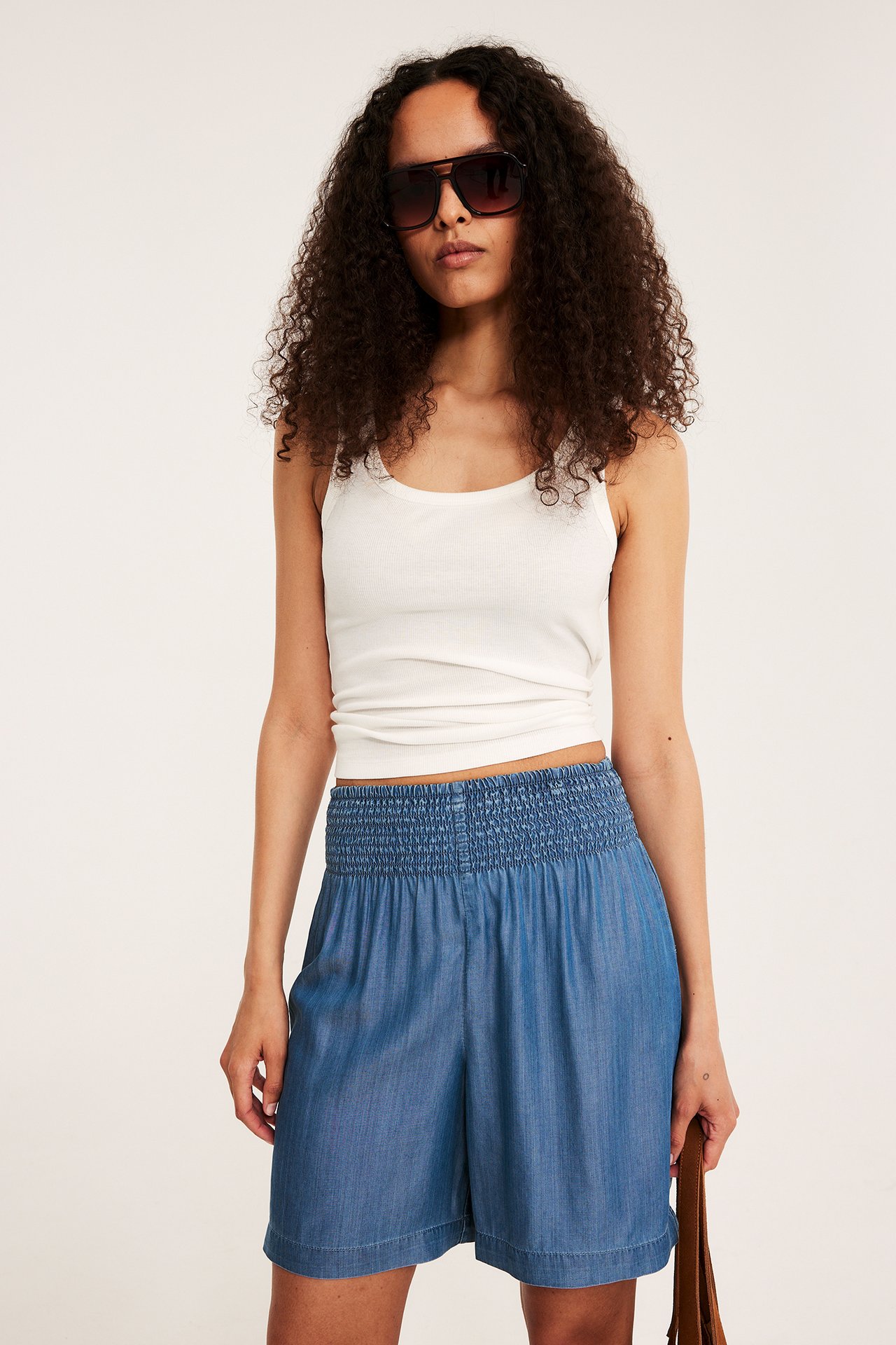 Widok z przodu: kobieta w niebieskich szortach Tencel z marszczoną talią, białym topie, ciemnych okularach.
