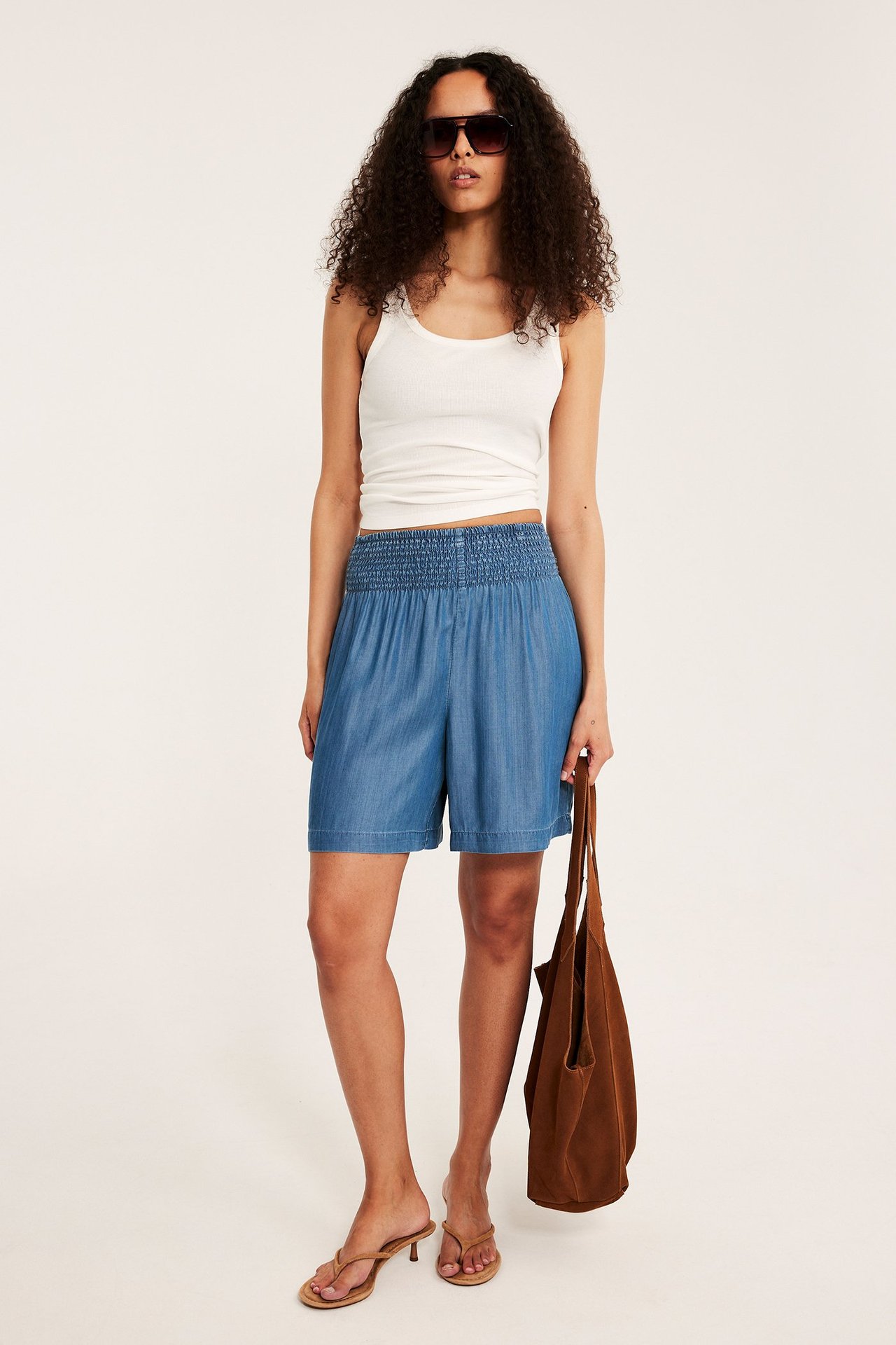 Kobieta nosi niebieskie szorty Tencel ze smockiem, biały top, okulary, sandały, trzyma brązową torbę.