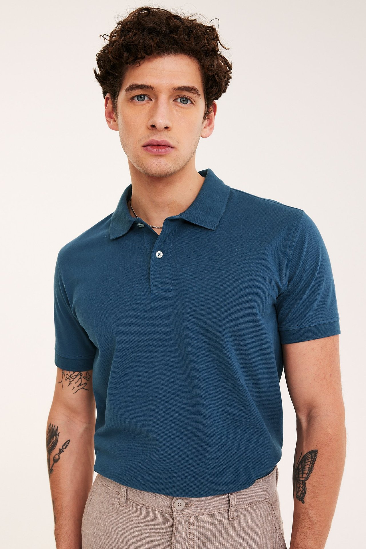 Framsida av en man i mörkblå bomullspikétröja med två vita knappar och ljusbruna byxor.