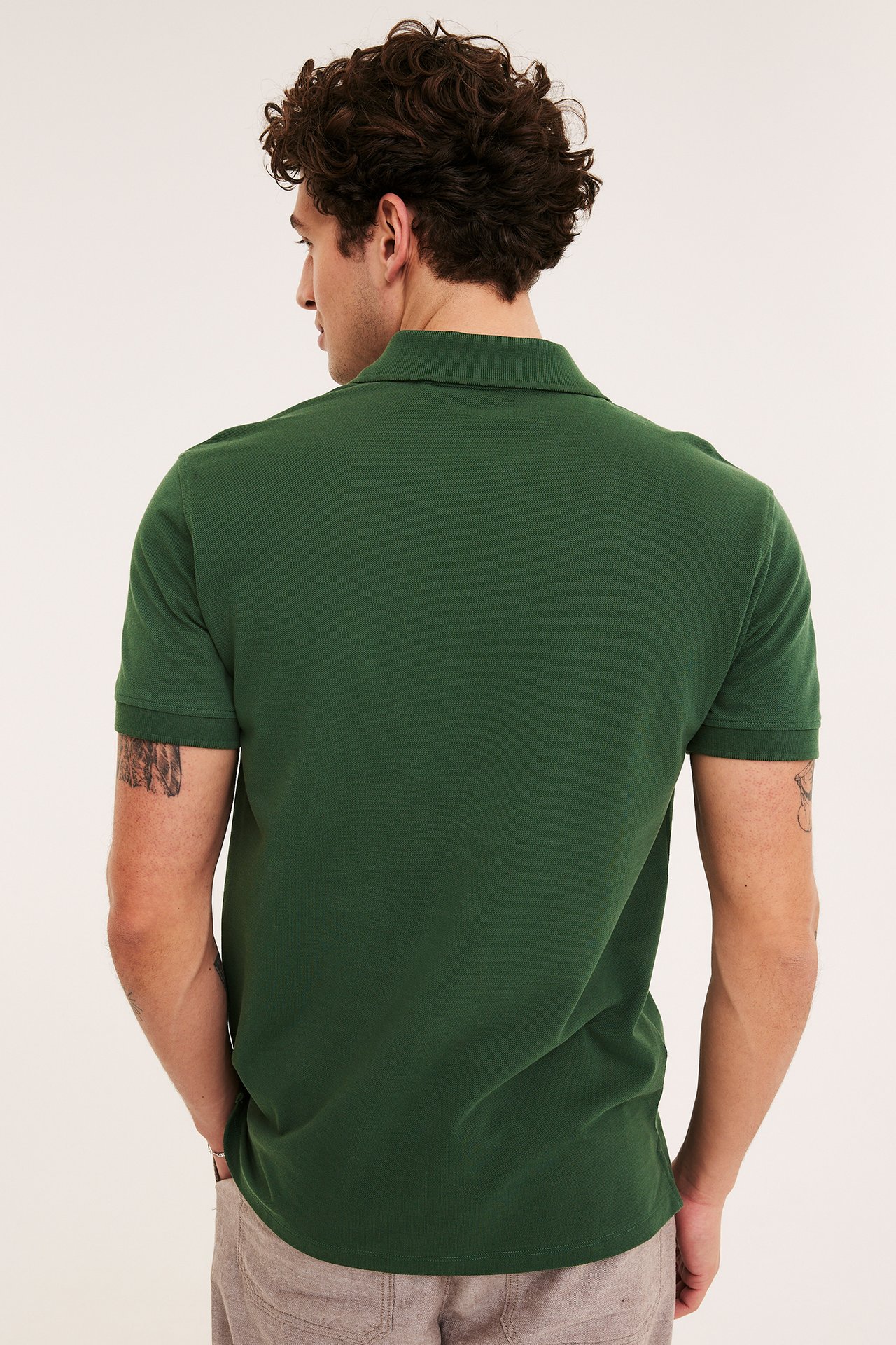 Bakifrån: En man i en enfärgad grön pikétröja av bomull och beiga byxor.