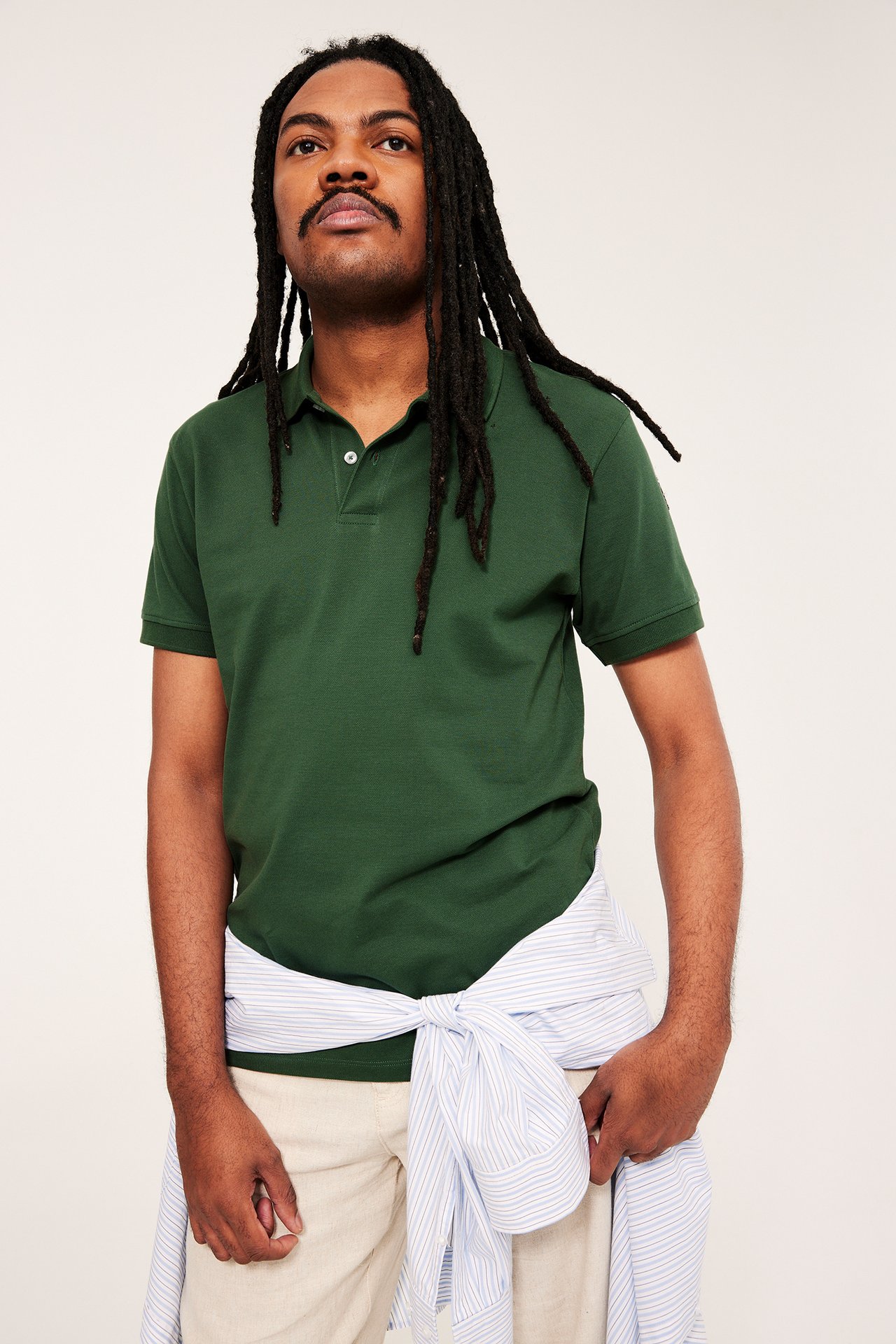 Mann forfra i grønn bomullspiké, beigefargede bukser og stripet skjorte knyttet rundt midjen.