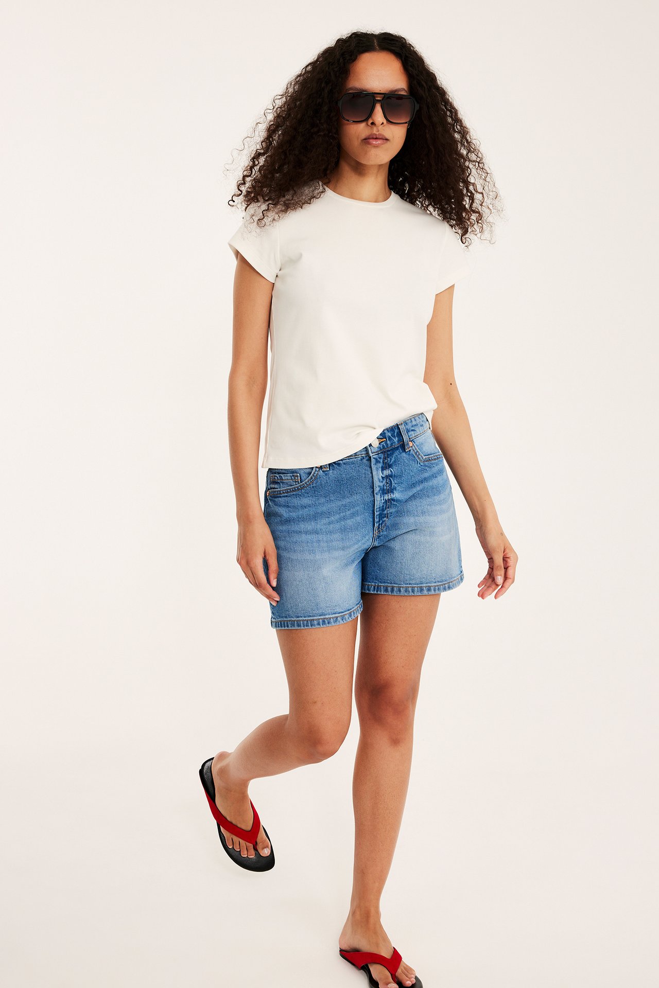 Edestäpäin nainen yllään vaaleansiniset denimshortsit, kermanvärinen t-paita, tummat aurinkolasit ja punaiset varvassandaalit.