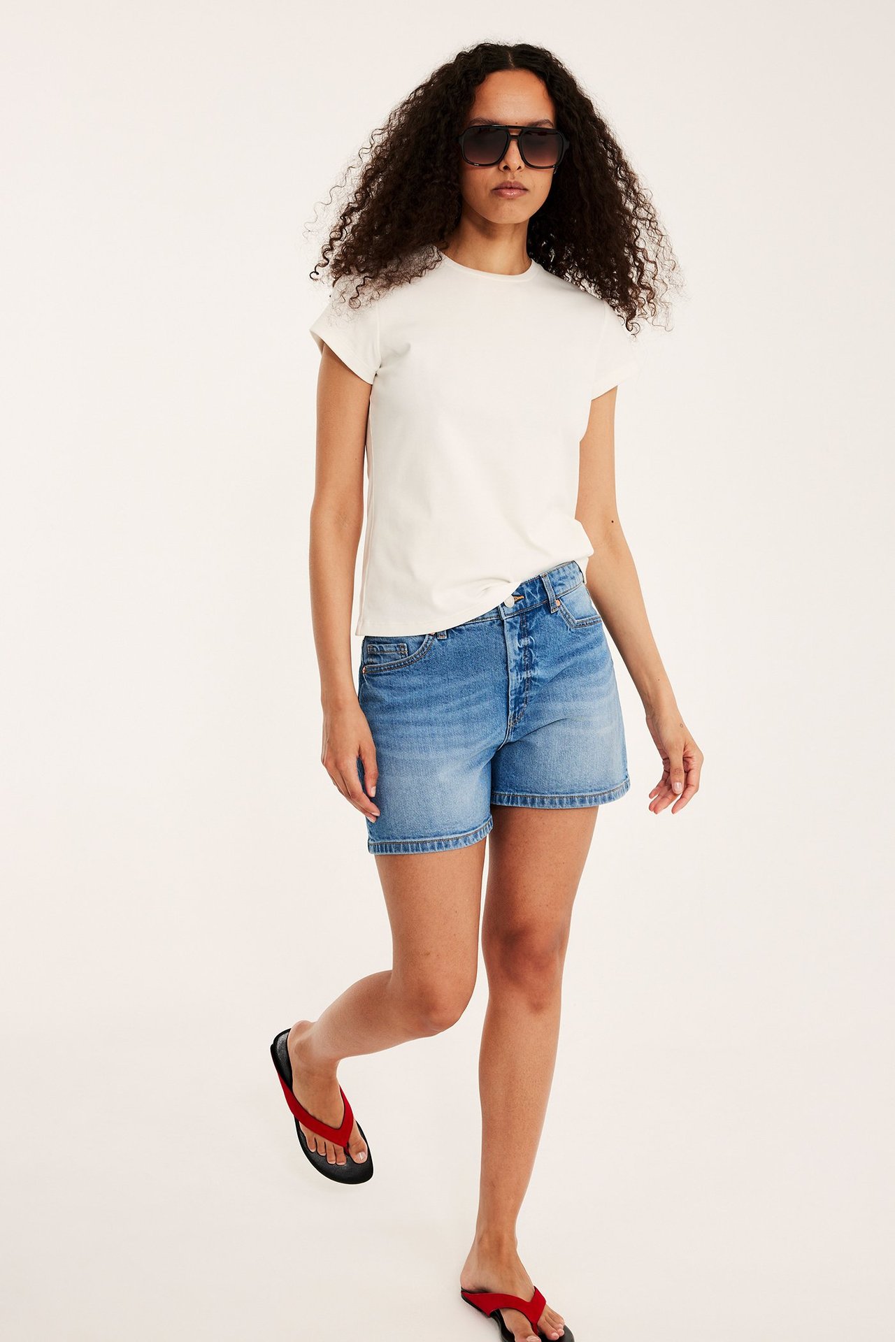 Edestäpäin nainen yllään vaaleansiniset denimshortsit, kermanvärinen t-paita, tummat aurinkolasit ja punaiset varvassandaalit.