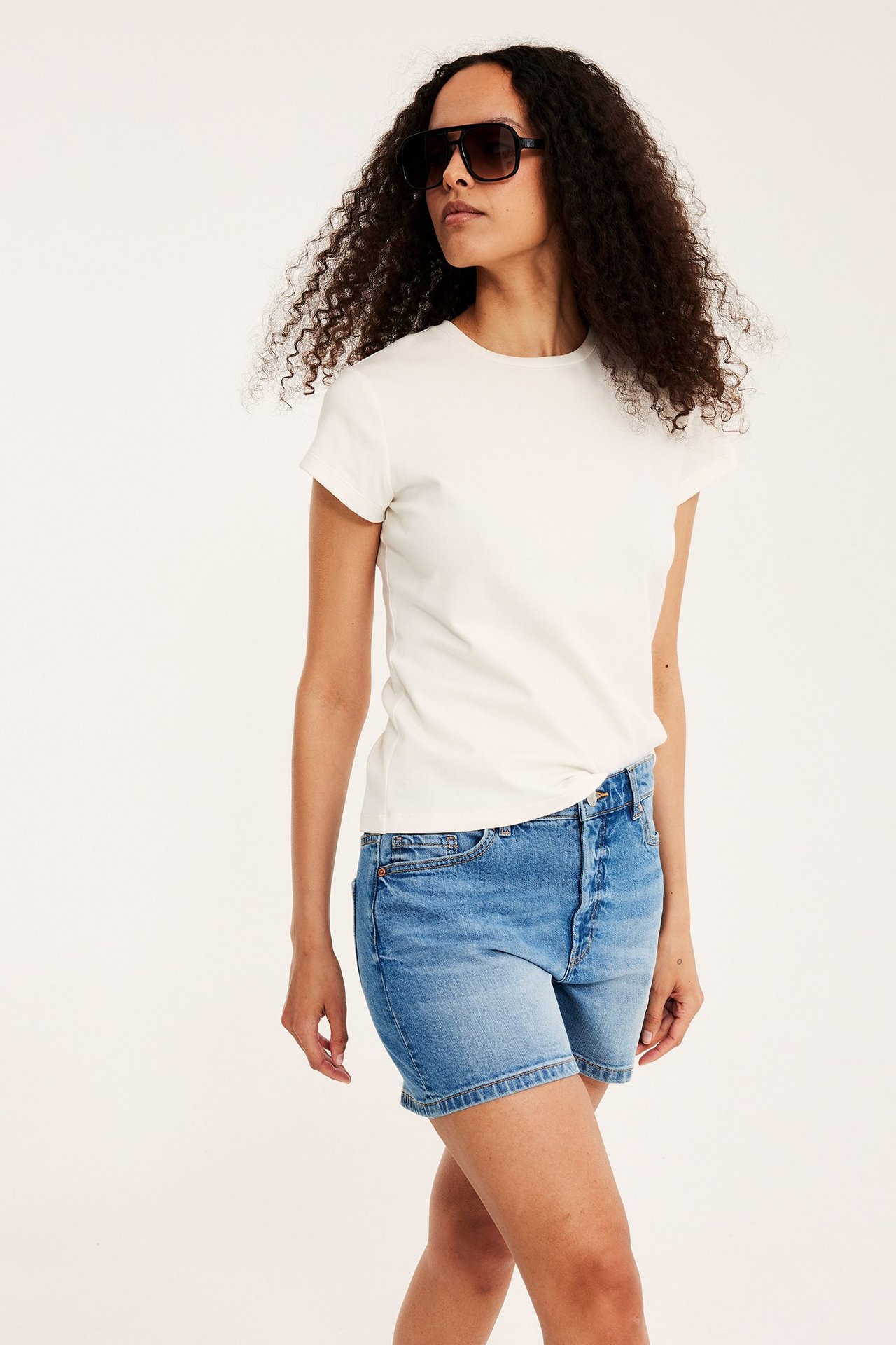 Edestä: nainen vaaleansinisissä denimshortseissa, valkoisessa t-paidassa ja tummissa aurinkolaseissa.