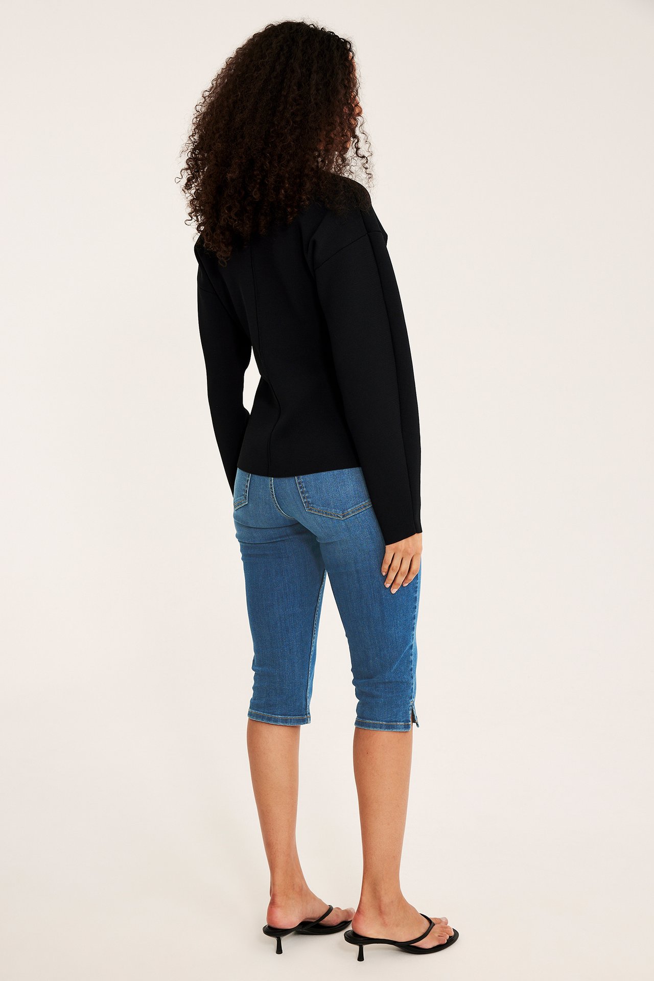 Widok z tyłu kobiety w niebieskich dżinsowych szortach capri, czarnym topie i czarnych sandałach na obcasie.