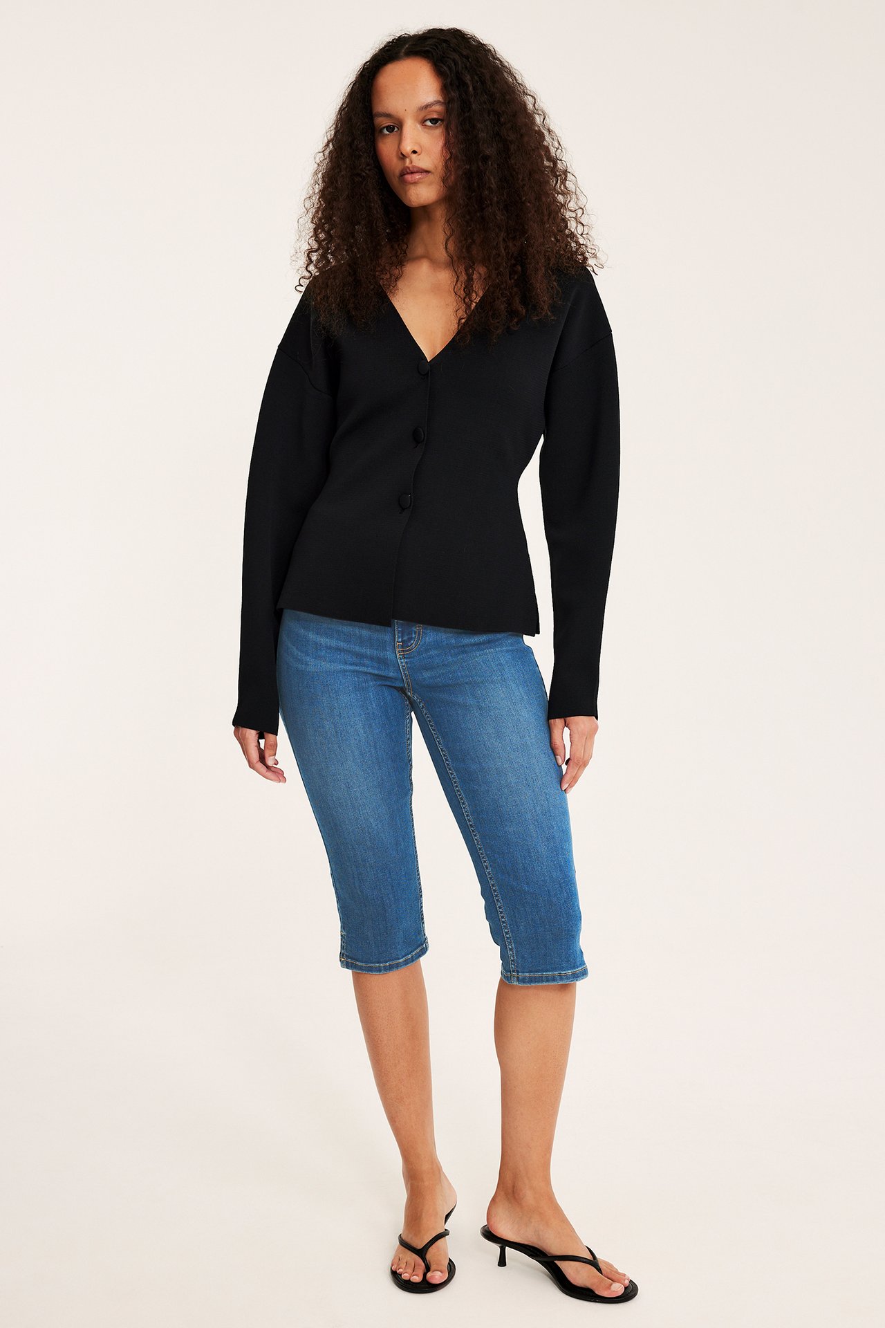 Forfra: kvinne i blå caprijeans, svart kneppet cardigan og svarte høyhælte sandaler.