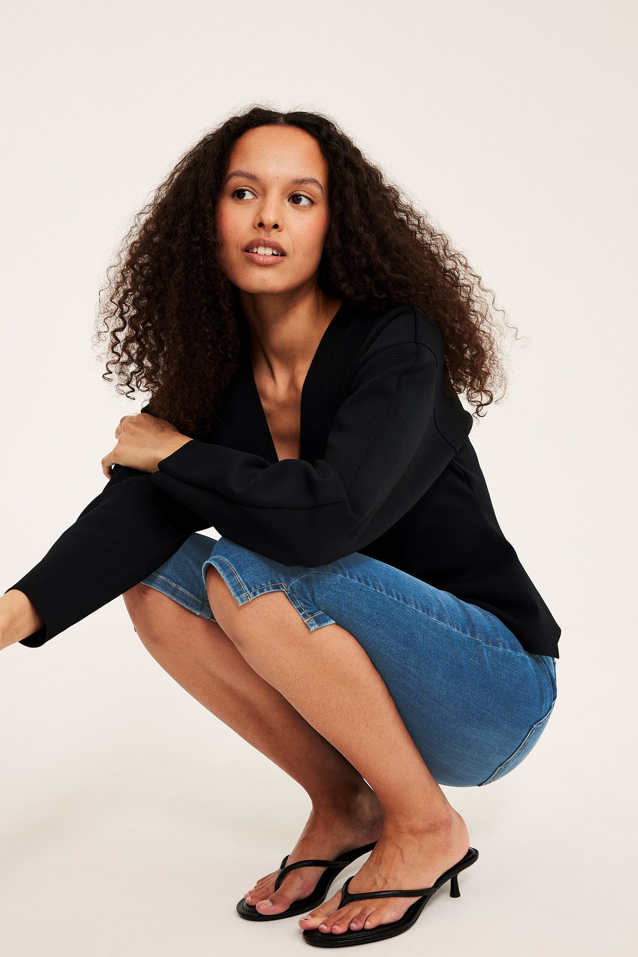 Side-frontvisning av en kvinne i blå denim capribukser, svart V-hals topp og svarte tåsandaletter.