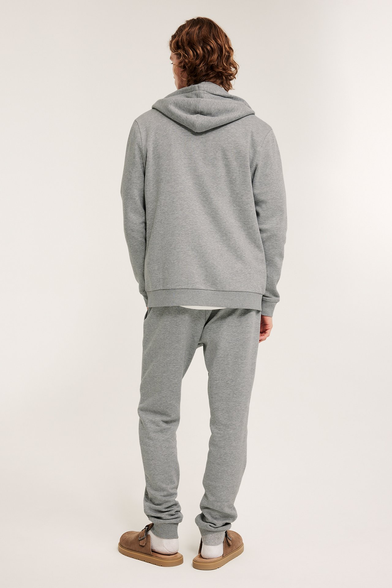 Takaapäin: mies vaaleanharmaissa collegehousuissa, joissa resorit, yhteensopiva huppari ja ruskeat tohvelit.