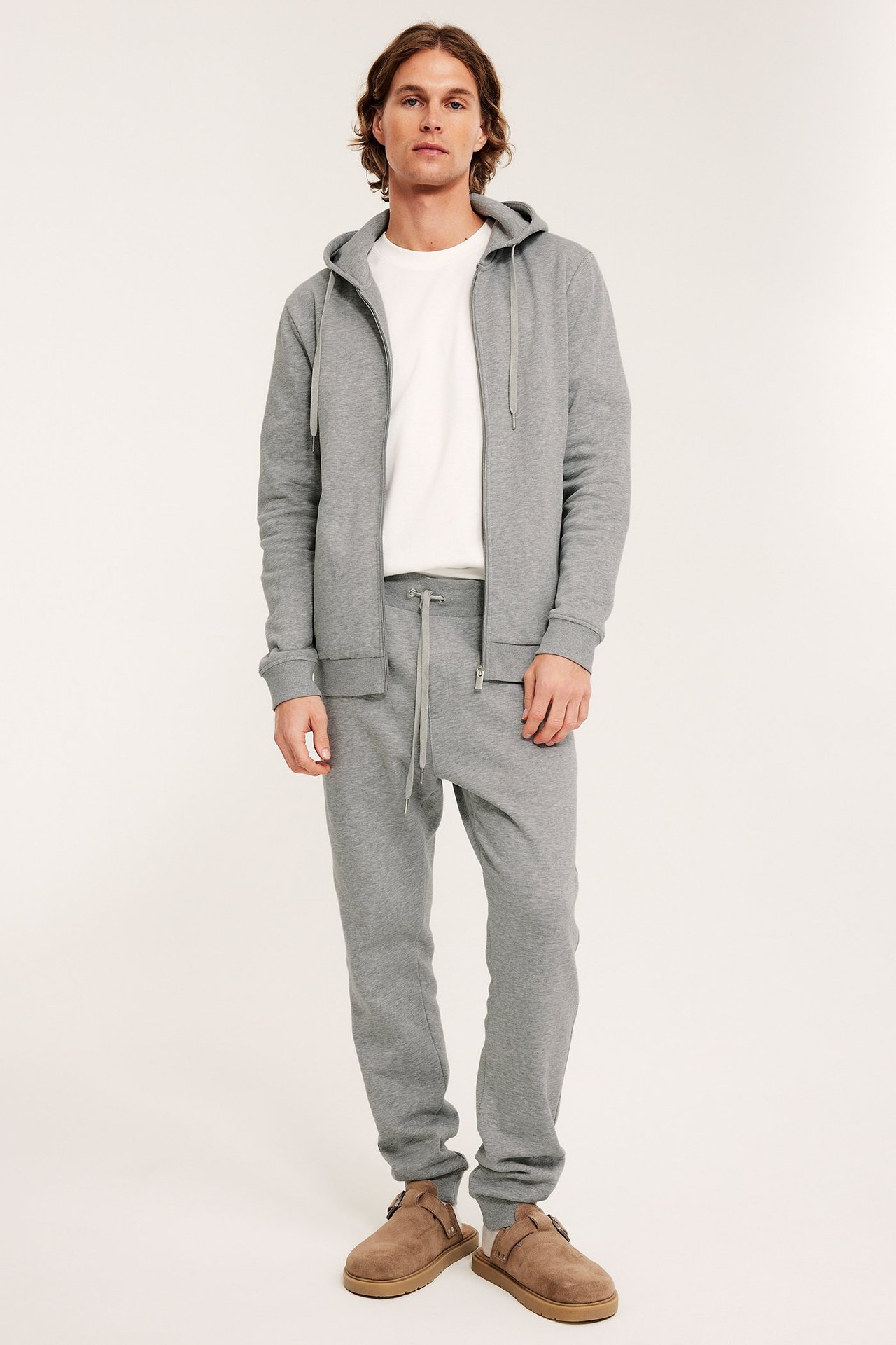 Man i ljusgrå melerade sweatpants, matchande huvtröja, vit t-shirt och bruna tofflor, framifrån.