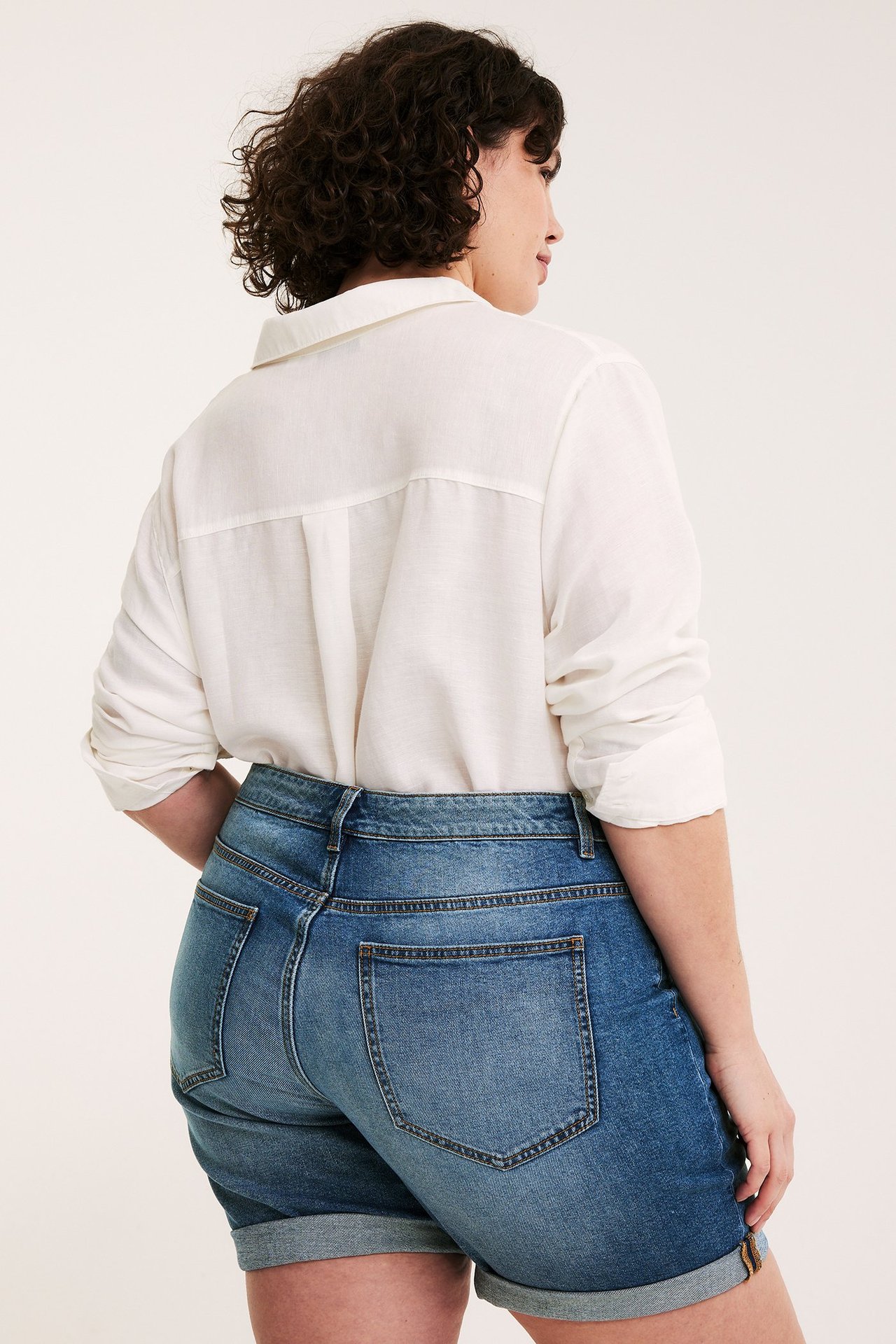 Naisen takakuva sinisissä farkkushortseissa, joissa on käärityt lahkeet, ja valkoisessa kauluspaidassa.