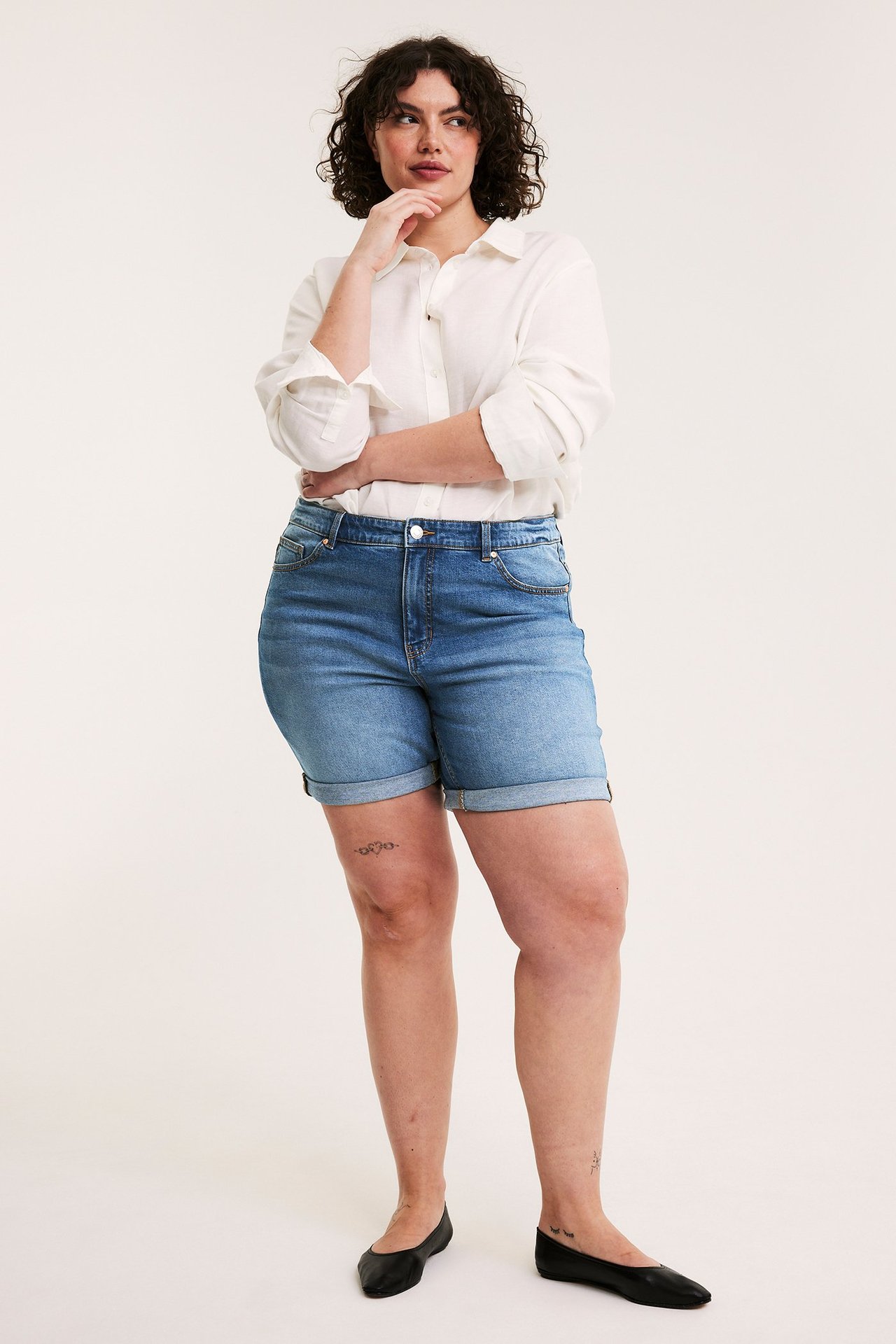 Nainen edestäpäin yllään siniset käärityt farkkushortsit, valkoinen paita ja mustat ballerinat.