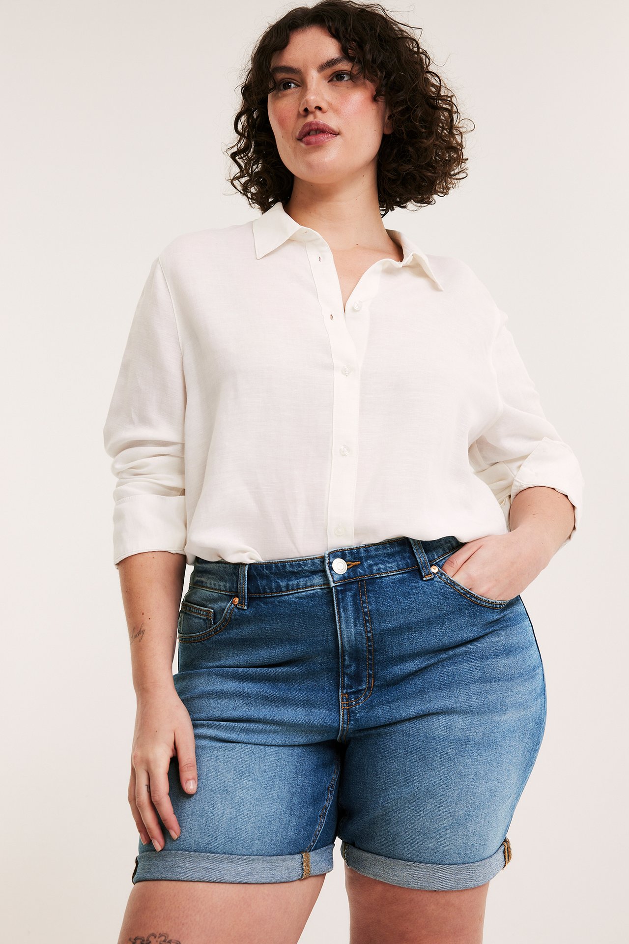 Nainen edestä päin yllään siniset denim-farkkushortsit käännetyillä lahkeilla ja valkoinen paitapusero.