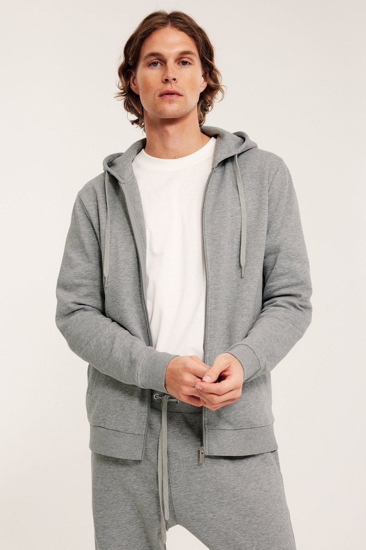 Mies edestä: vaaleanharmaa vetoketjuhuppari, valkoinen t-paita ja harmaat collegehousut.