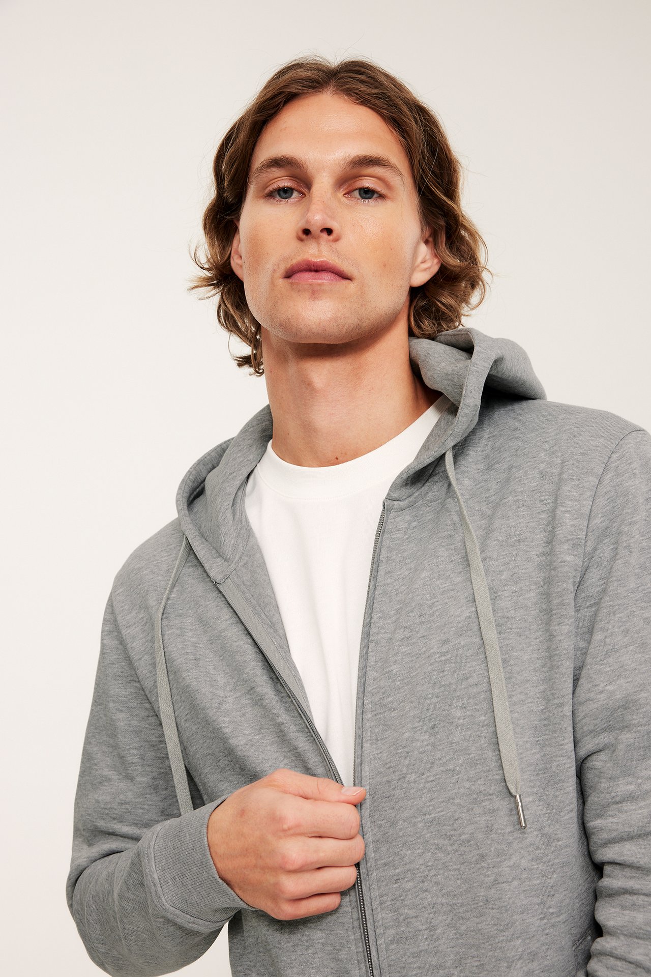 Lähikuva edestä: mies yllään vaaleanharmaa meleerattu vetoketjuhuppari valkoisen t-paidan päällä.