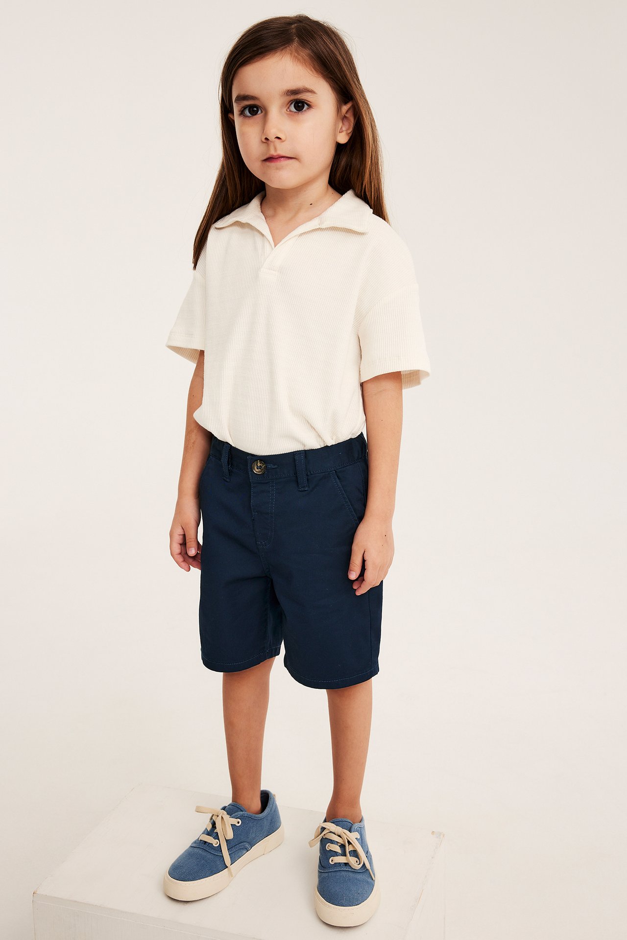 Widok z przodu: dziecko w granatowych bawełnianych szortach chinos, kremowej koszulce polo i niebieskich trampkach.