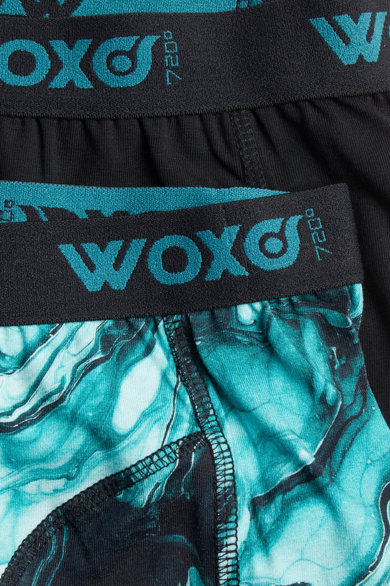 Närbild på två par boxerkalsonger för barn: en mönstrad grön/svart och en svart, med teal WOXO-resår.