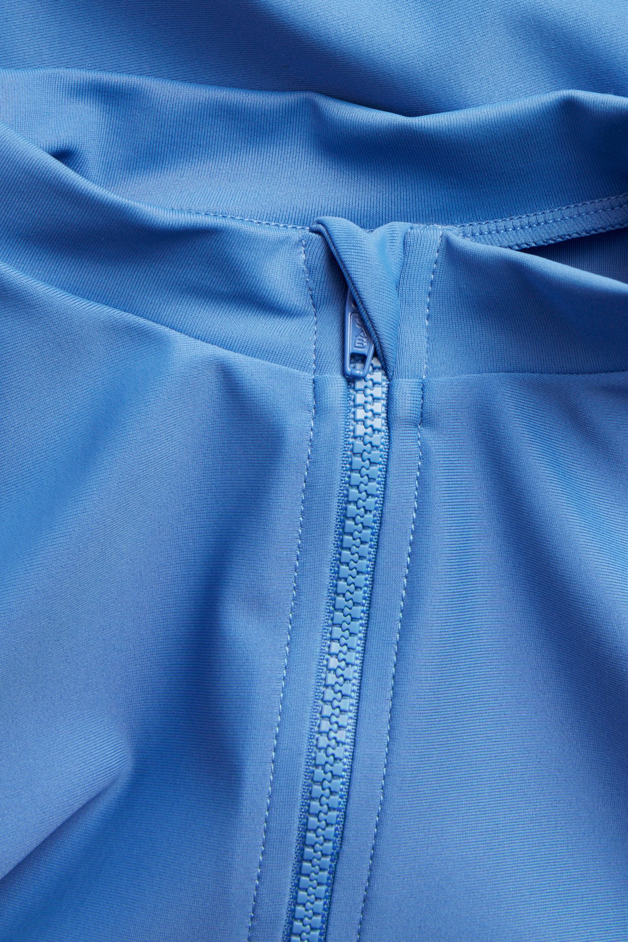 Nærbilde av en blå ensfarget langermet UV-topp i polyester med matchende blå glidelås.