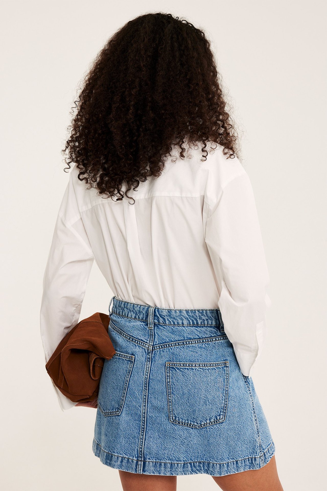 Bakifrån: kvinna i ljusblå A-linjeformad jeanskjol och vit skjorta, håller en brun sak.