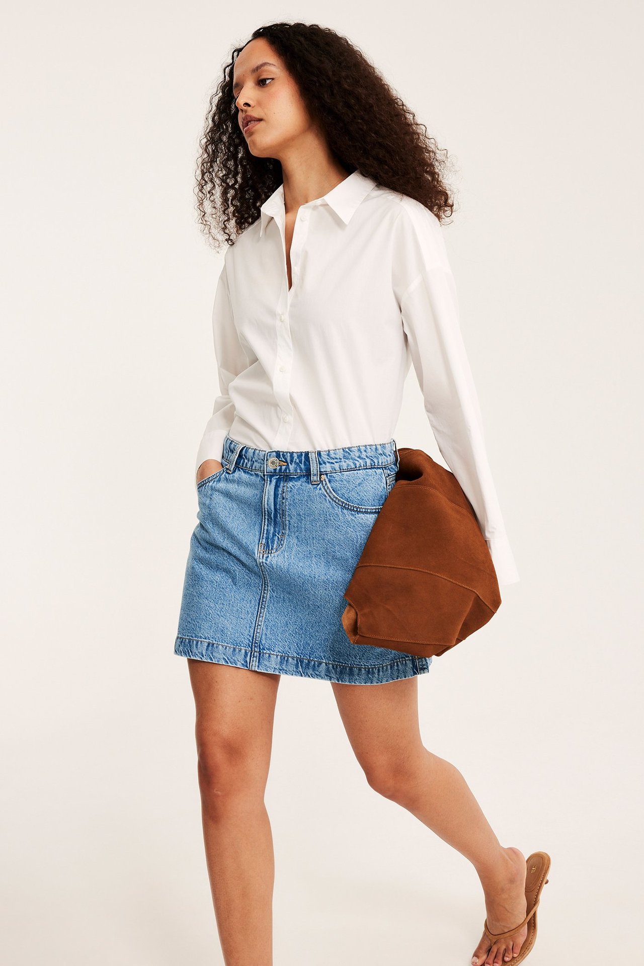 Edestäpäin: nainen vaaleansinisessä A-linjaisessa farkkuhameessa, valkoisessa paidassa, pitää ruskeaa laukkua.