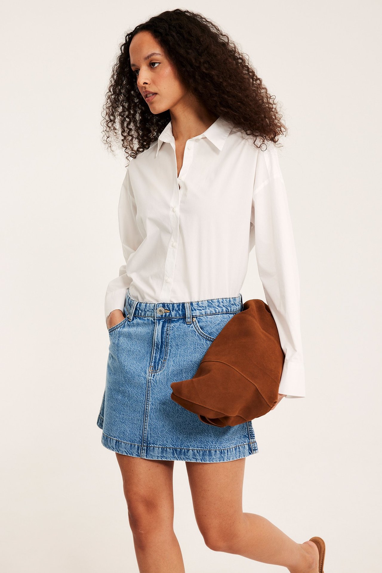 Kvinna i ljusblå A-linjeformad jeanskjol, vit skjorta, håller en brun clutchväska.