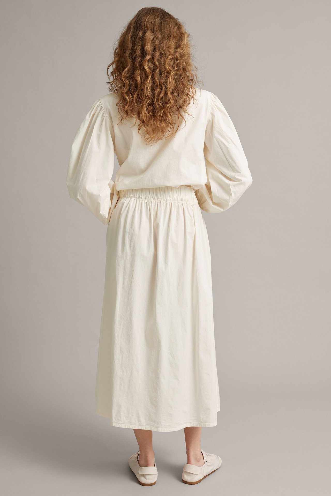 Takakuva: Nainen valkoisessa popliinikietaimihameessa joustovyötäröllä ja vaaleassa puserossa.