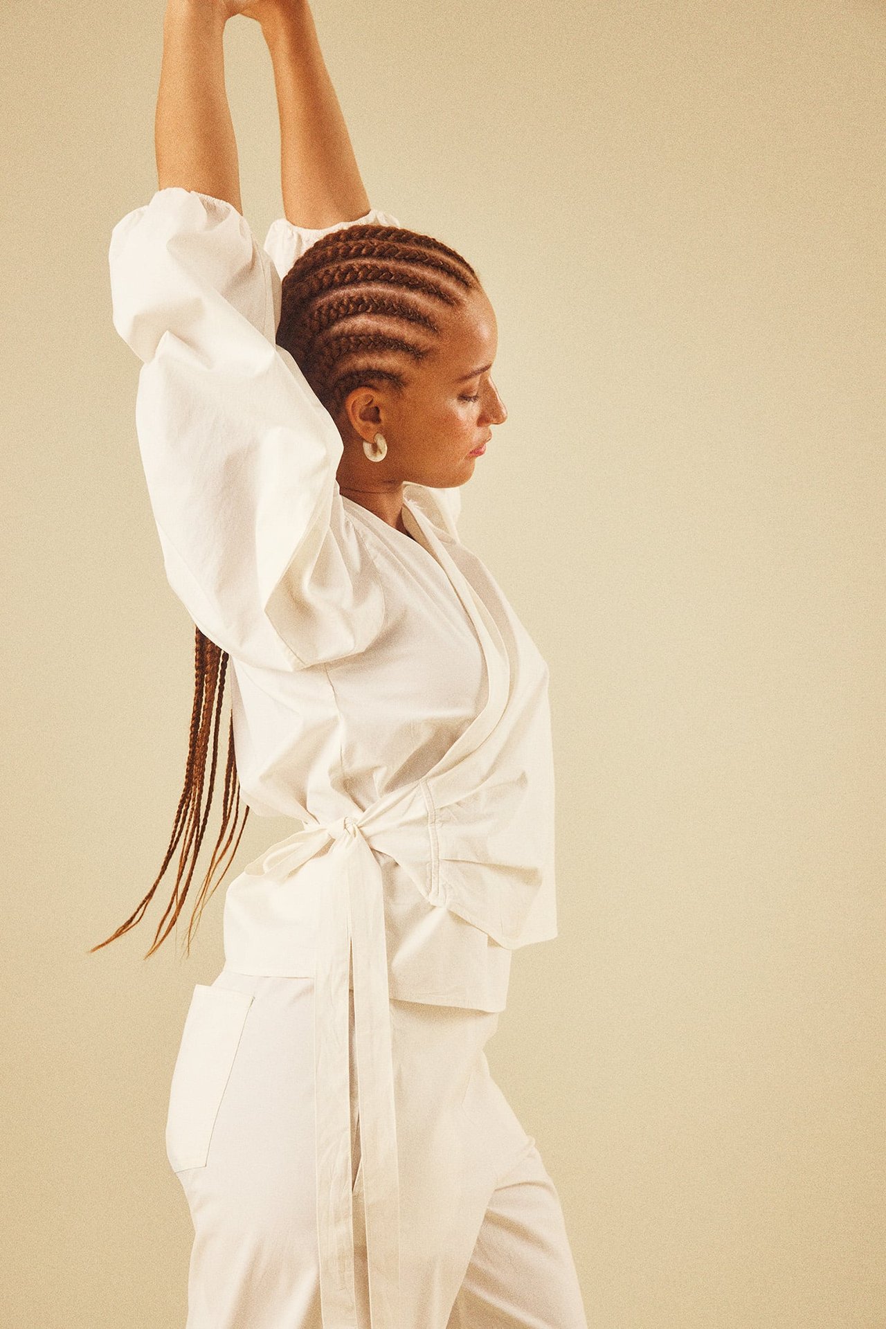 Sidovy av kvinna i vit omlott-topp med voluminösa ärmar och knytning, matchande vita byxor.