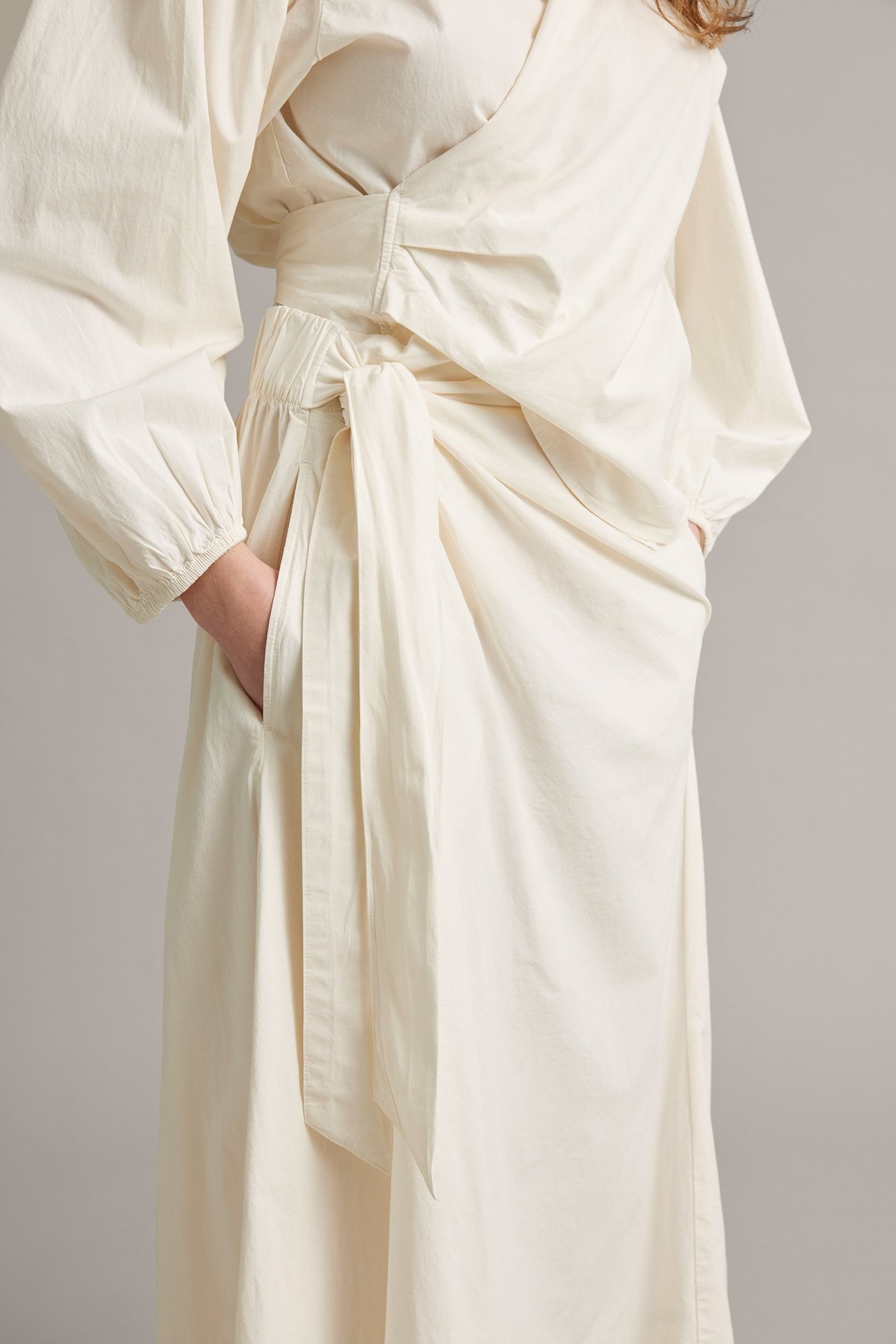 Nærbilde forfra: kvinne i hvitt poplin omslagsskjørt med knyting, stylet med matchende topp, hånd i lommen.