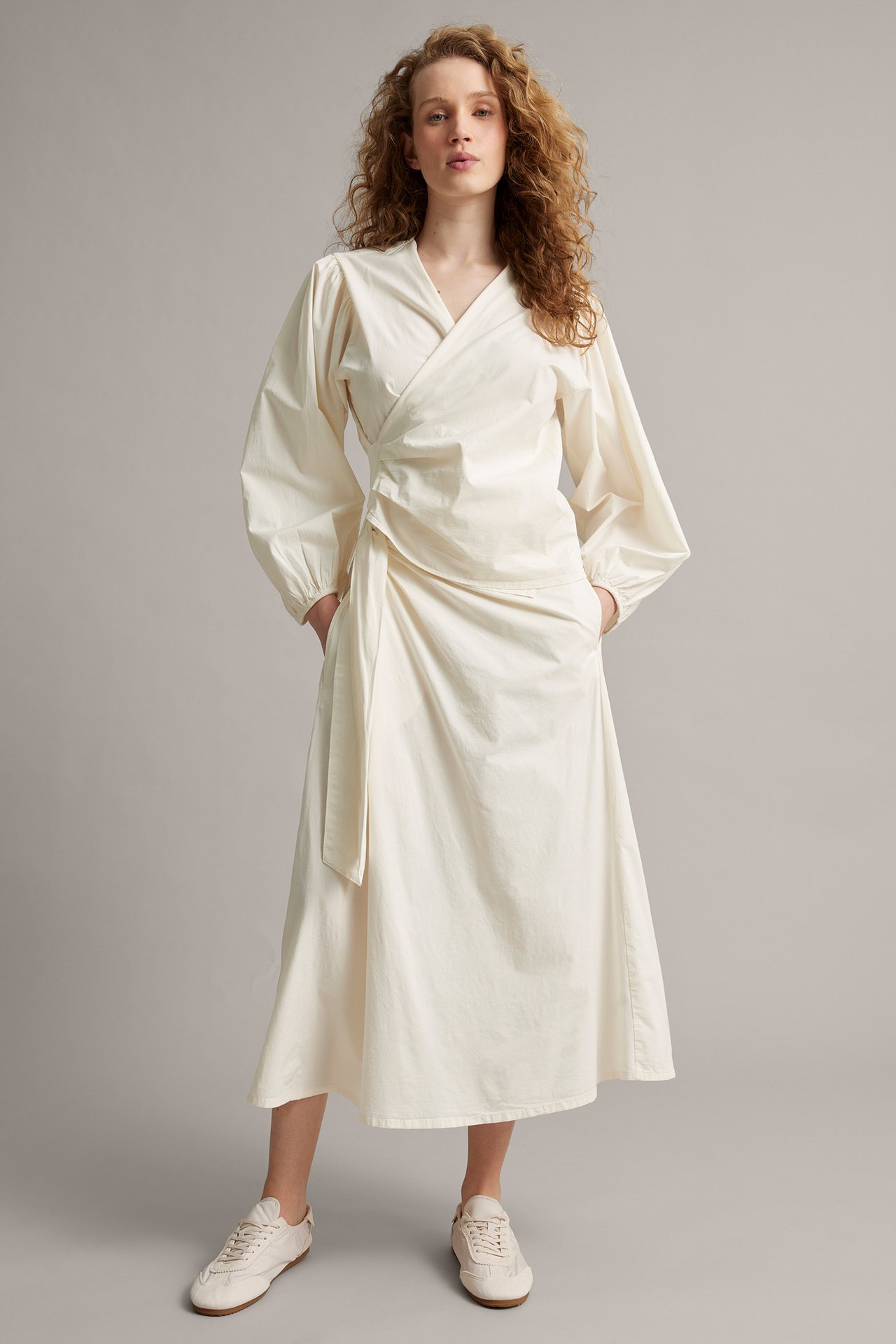 Kobieta w białej, popelinowej sukience kopertowej z wiązaniem z boku i jasnymi sneakersami, widok z przodu.