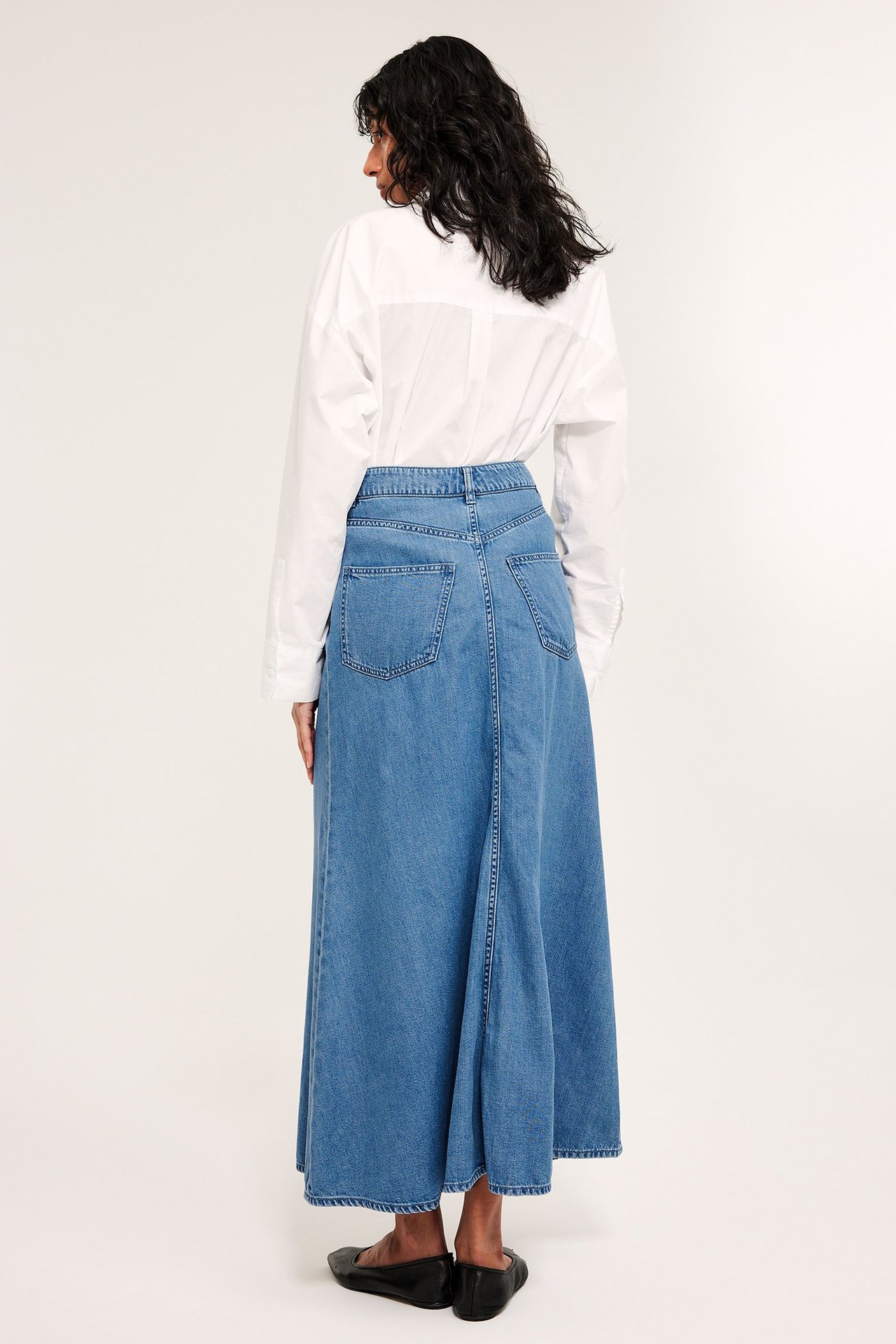 Ryggvy av kvinna i lång utsvängd blå jeanskjol, vit långärmad skjorta och svarta ballerinaskor.