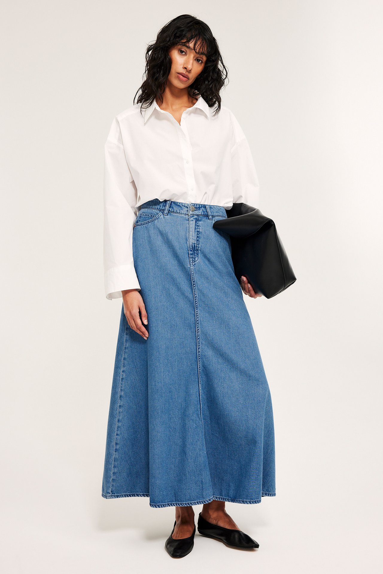 Frontvy: Kvinna i lång, utsvängd denimblå kjol, vit skjorta, svarta ballerinaskor, håller svart kuvertväska.