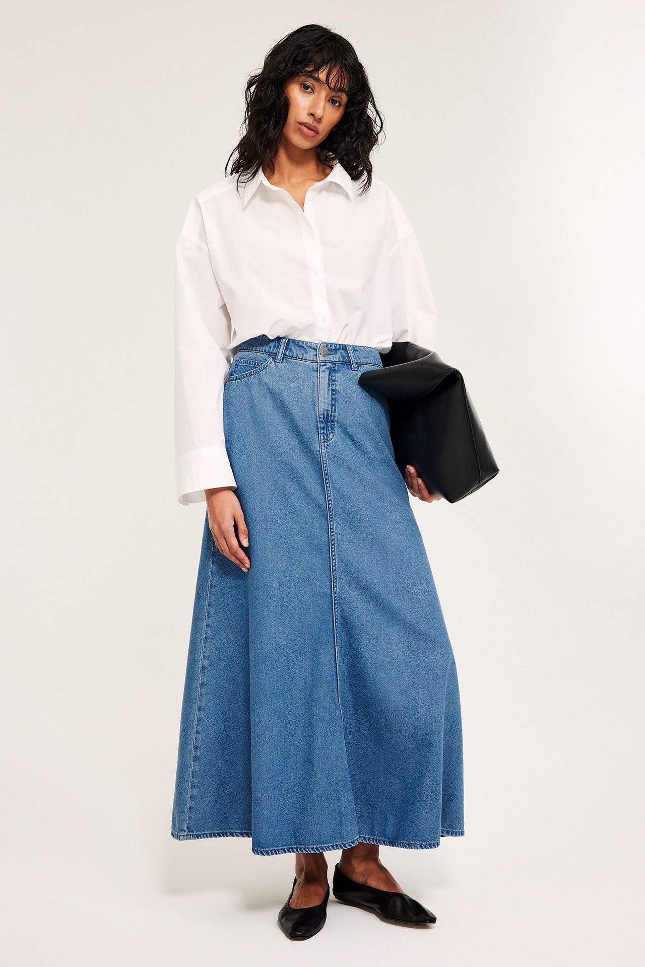 Frontvy: Kvinna i lång, utsvängd denimblå kjol, vit skjorta, svarta ballerinaskor, håller svart kuvertväska.