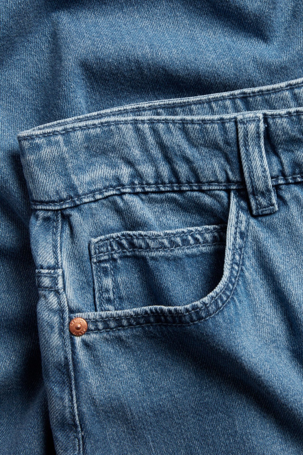 Zbliżenie z przodu niebieskiej spódnicy jeansowej z kieszenią, miedzianym nitem i szlufkami.