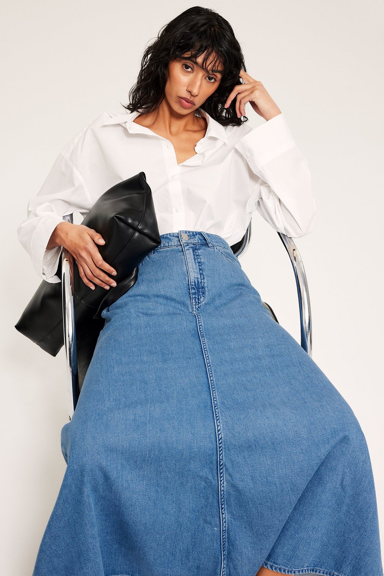 Framifrån: Kvinna i klockad blå jeanskjol och vit skjorta, håller en svart vadderad handväska.