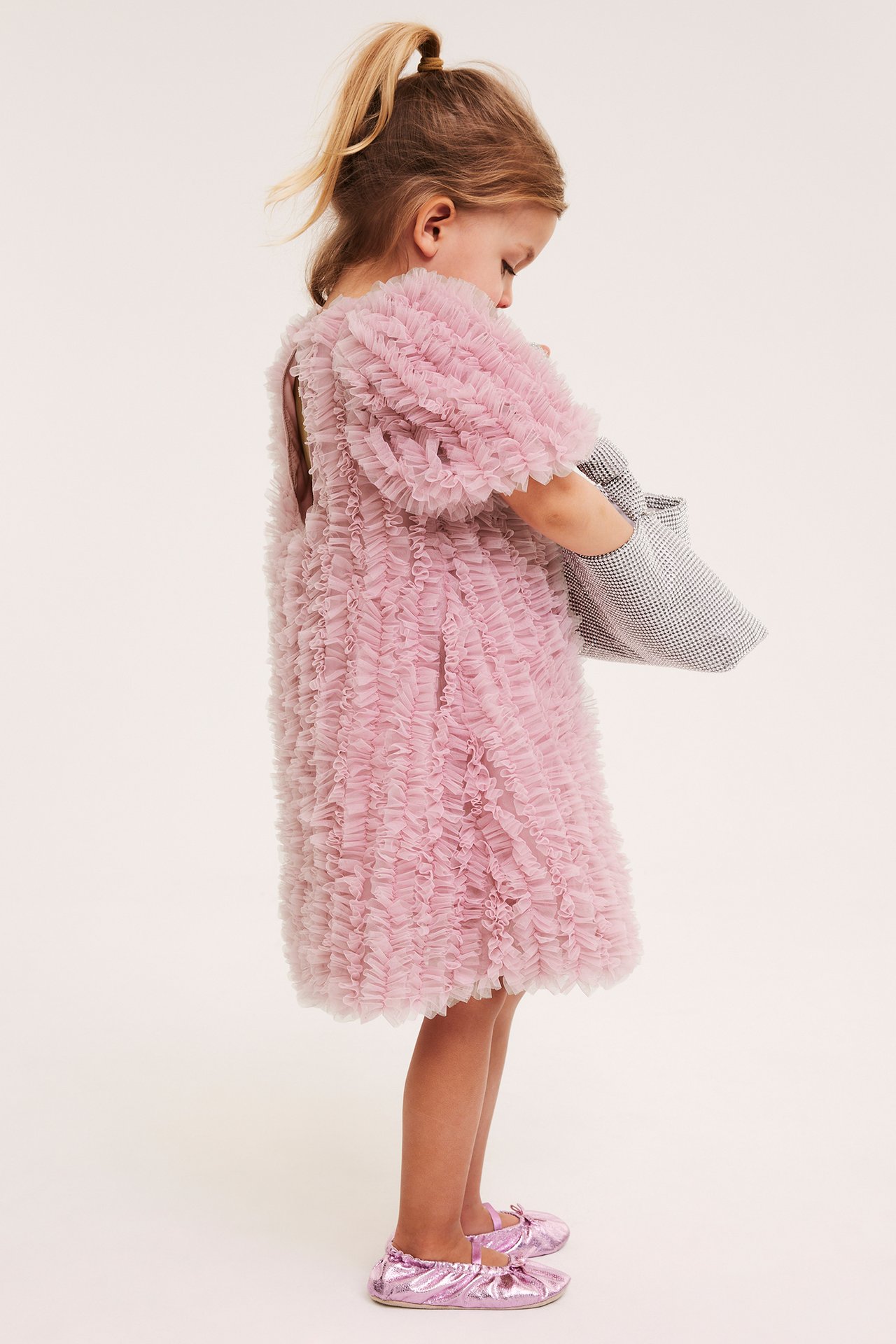 Sidebilde av et barn i lys rosa rynket meshkjole, holdende en rutete veske og metallrosa ballettsko.