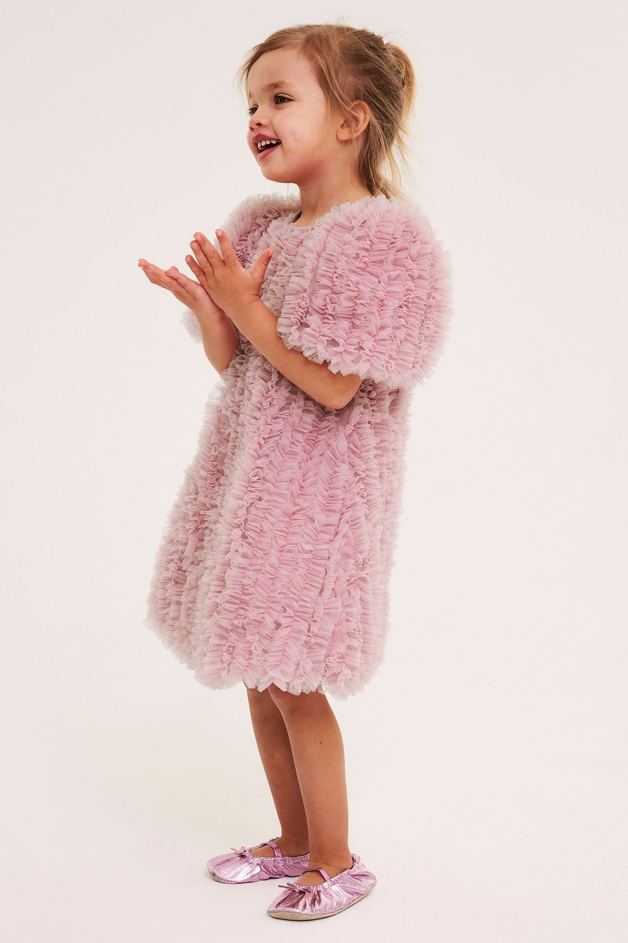 Sivukuva taputtavasta lapsesta, yllään vaaleanpunainen A-linjainen rypytetty verkkomekko ja metallinvaaleanpunaiset balettitossut.