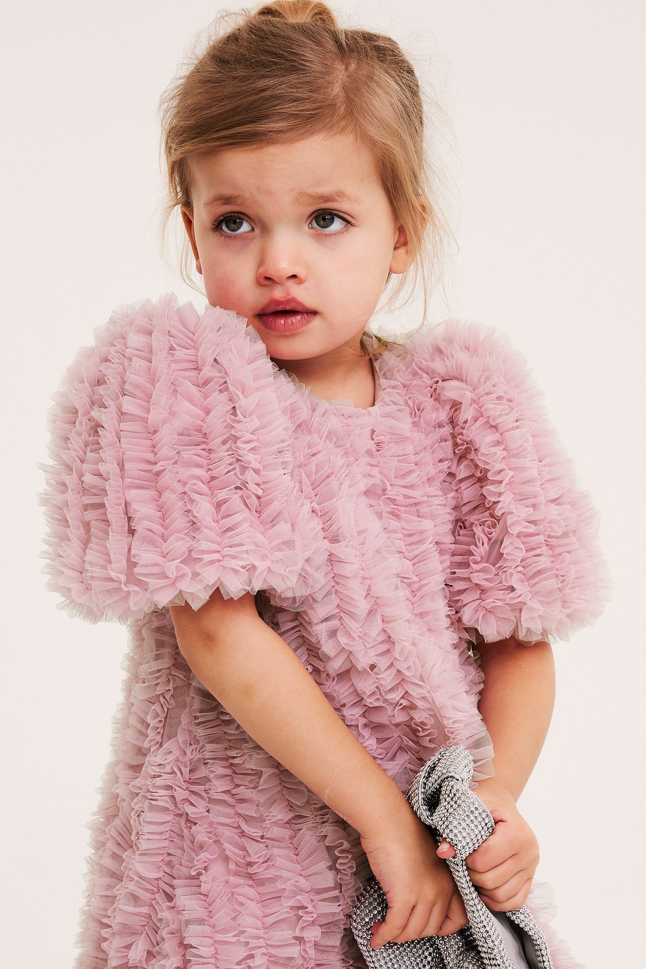Z przodu, dziecko w jasnoróżowej sukience z falbanami z siateczki, trzyma srebrną torebkę z cekinami.