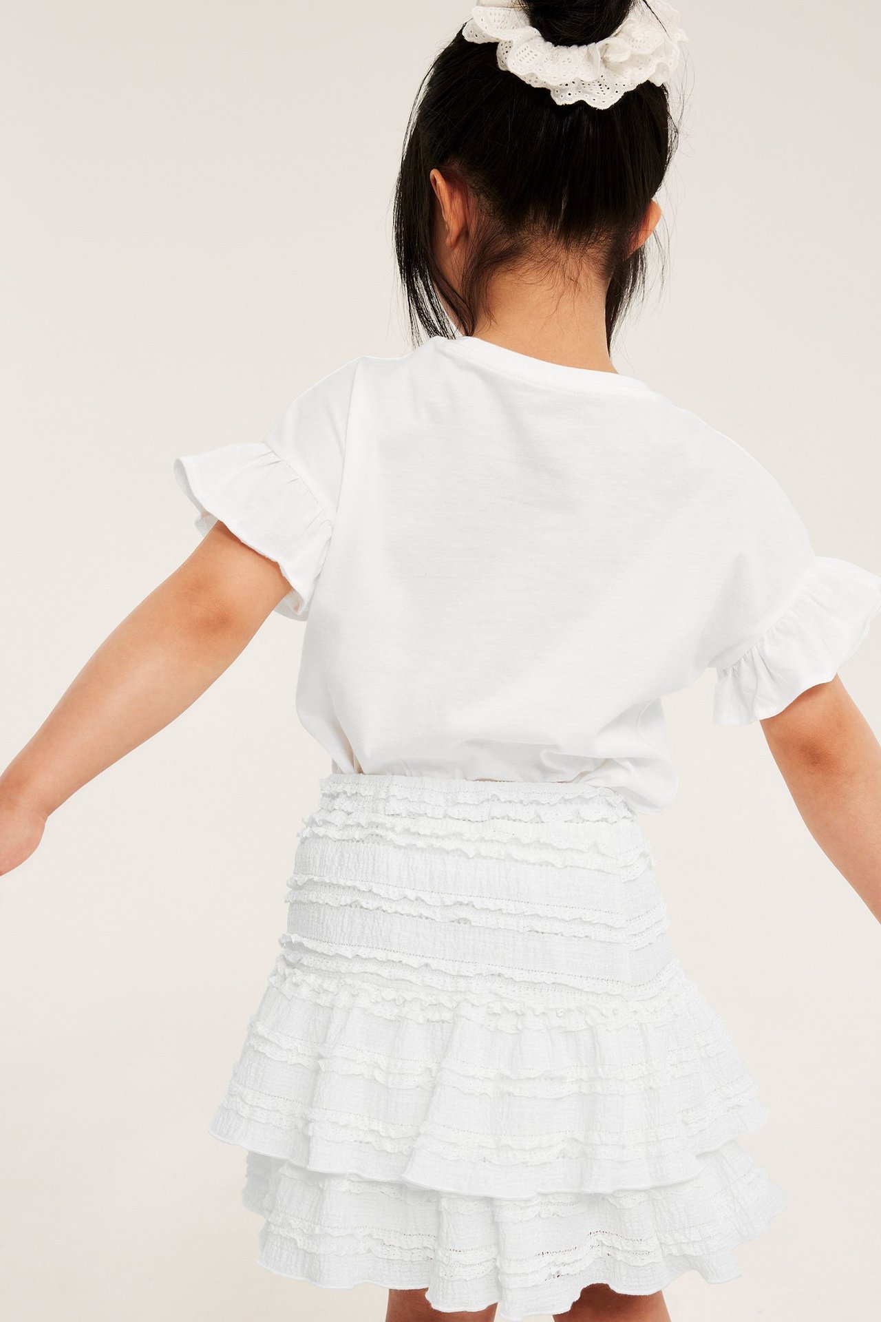 Bakifrån: kid i offwhite volangspetskjol med resår, vit t-shirt med volanger och vit spetsscrunchie i håret.