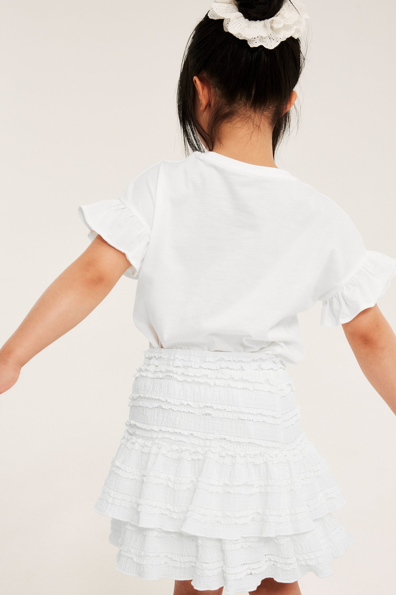 Bakifrån: kid i offwhite volangspetskjol med resår, vit t-shirt med volanger och vit spetsscrunchie i håret.