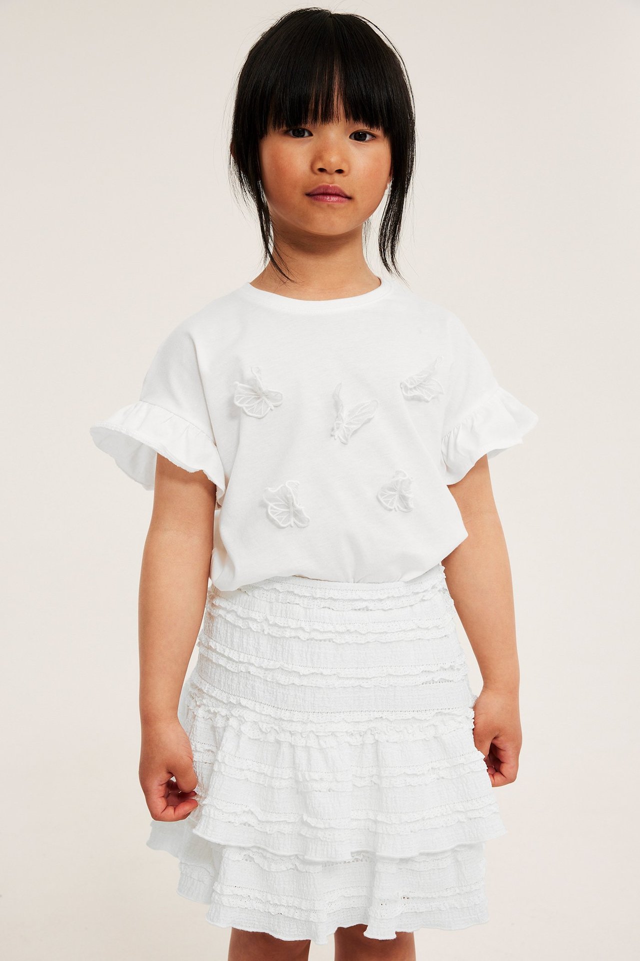Frontvy av ett barn i en offwhite volangspetskjol med resår i midjan och en vit t-shirt.